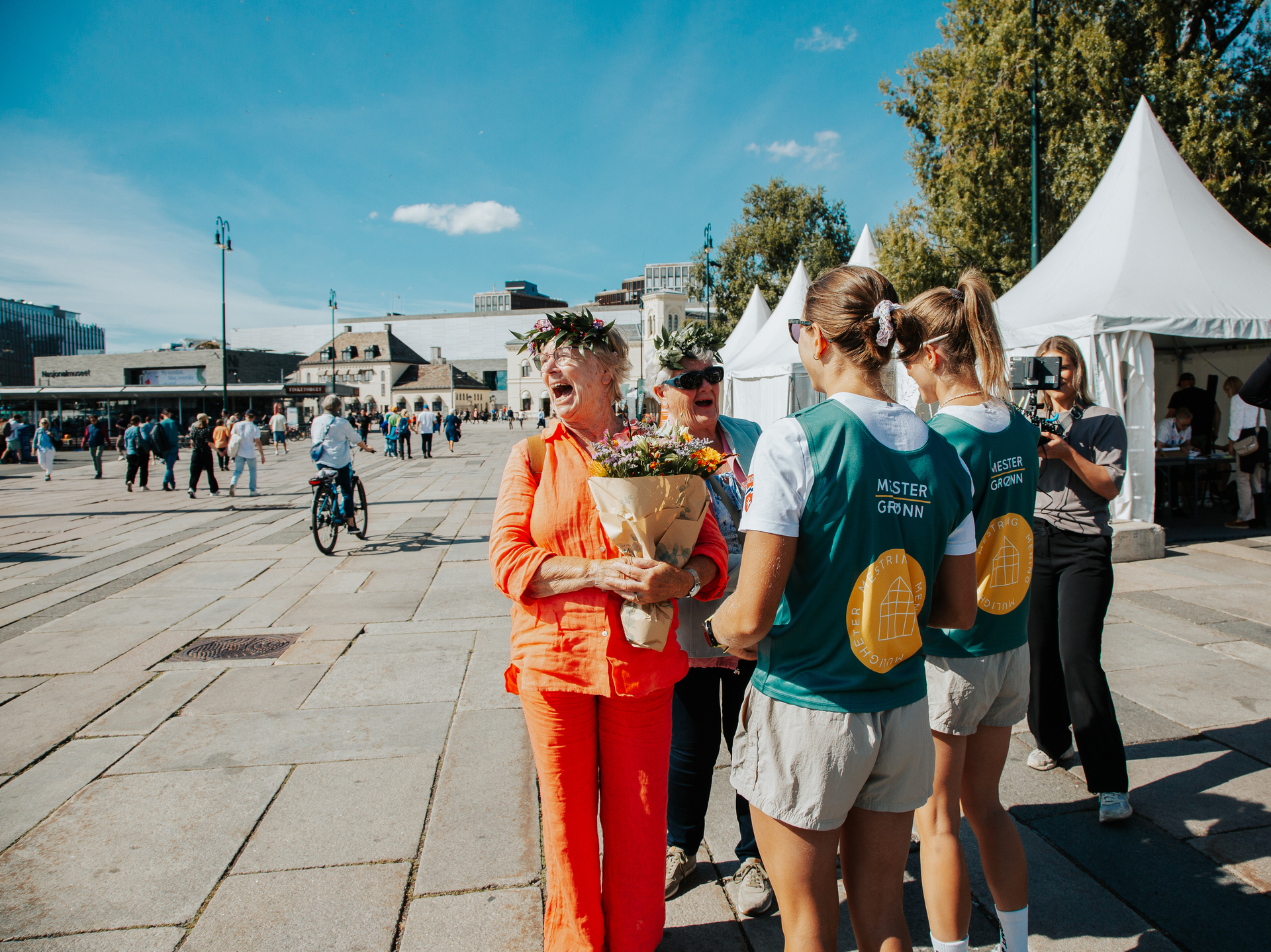 Foto: Kristin Austgulen Fosnæs og Nora Sannes står med ryggen mot kamera, i mester grønn vester. Foran dem står to eldre kvinner med blomster i håret og ser ut som de ler. Den ene kvinnen har oransje bluse og bukse og holder en stor blomsterbukett. den andre har på seg turkis jakke og solbriller.