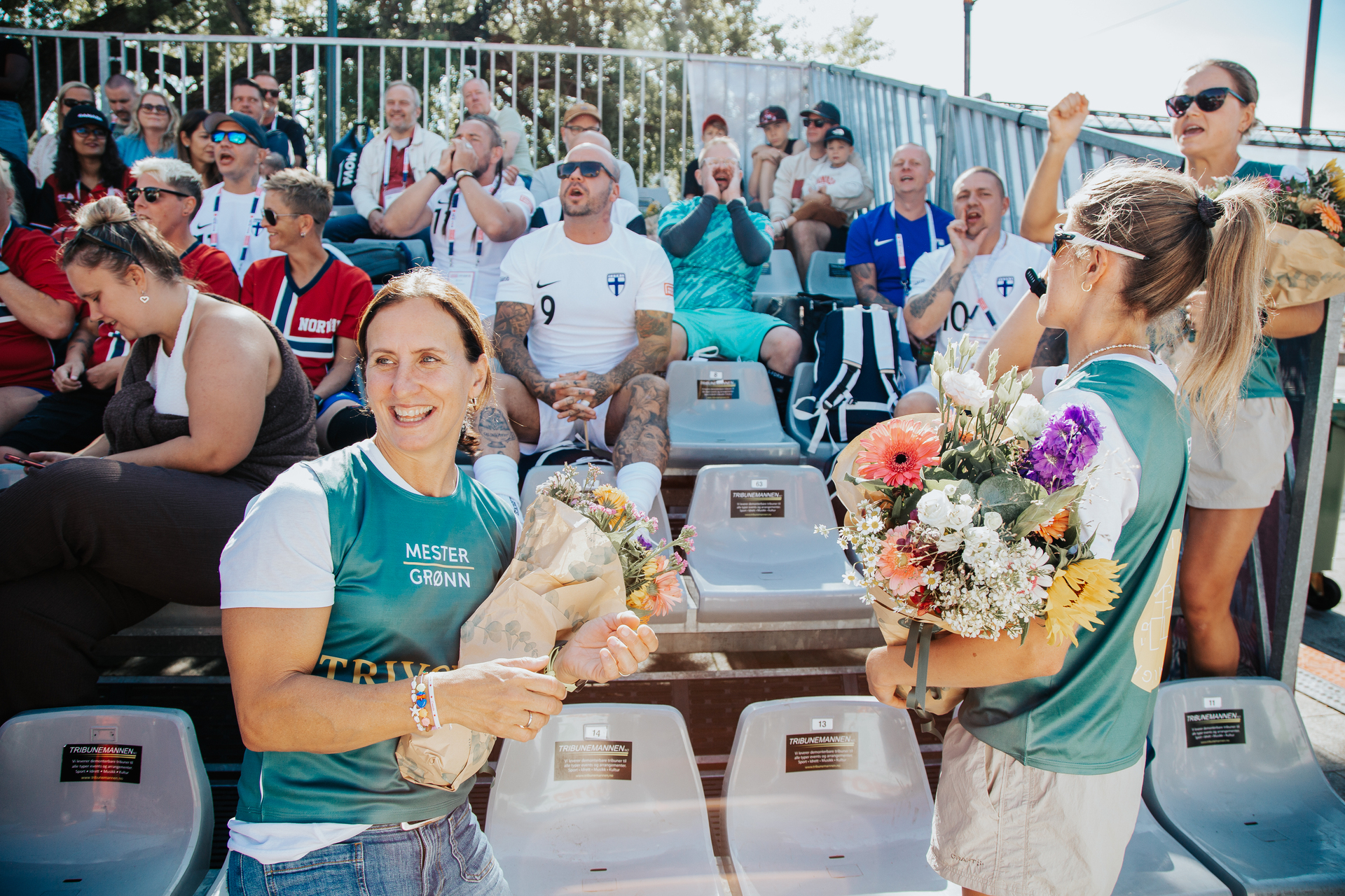 Foto: Marit Bjørgen og Kristin Austgulen Fosnæs står på tribunen under en fotballkamp, de har på seg grønne mester grønn-vester. Bjørgen ser mot banen og klapper, Fosnæs står med siden mot kamera og smiler. Bak dem sees mange publikummere som roper og heier.