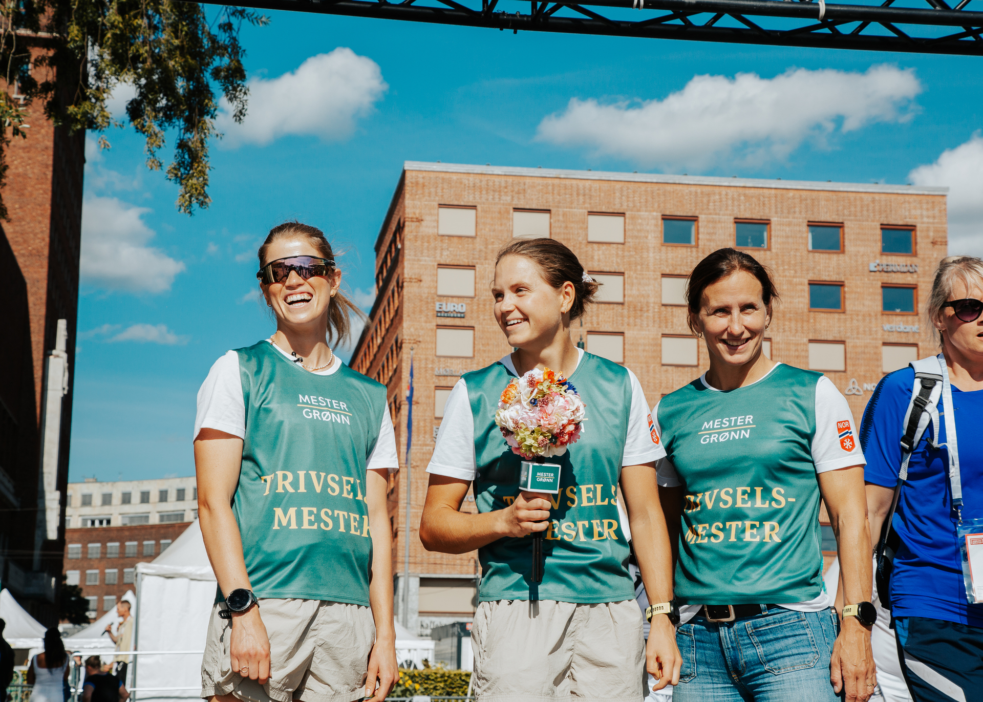 Marit Bjørgen står sammen med to andre langrennsjenter. Alle tre har på seg grønn vest fra mester grønn. Alle tre smiler mot siden.