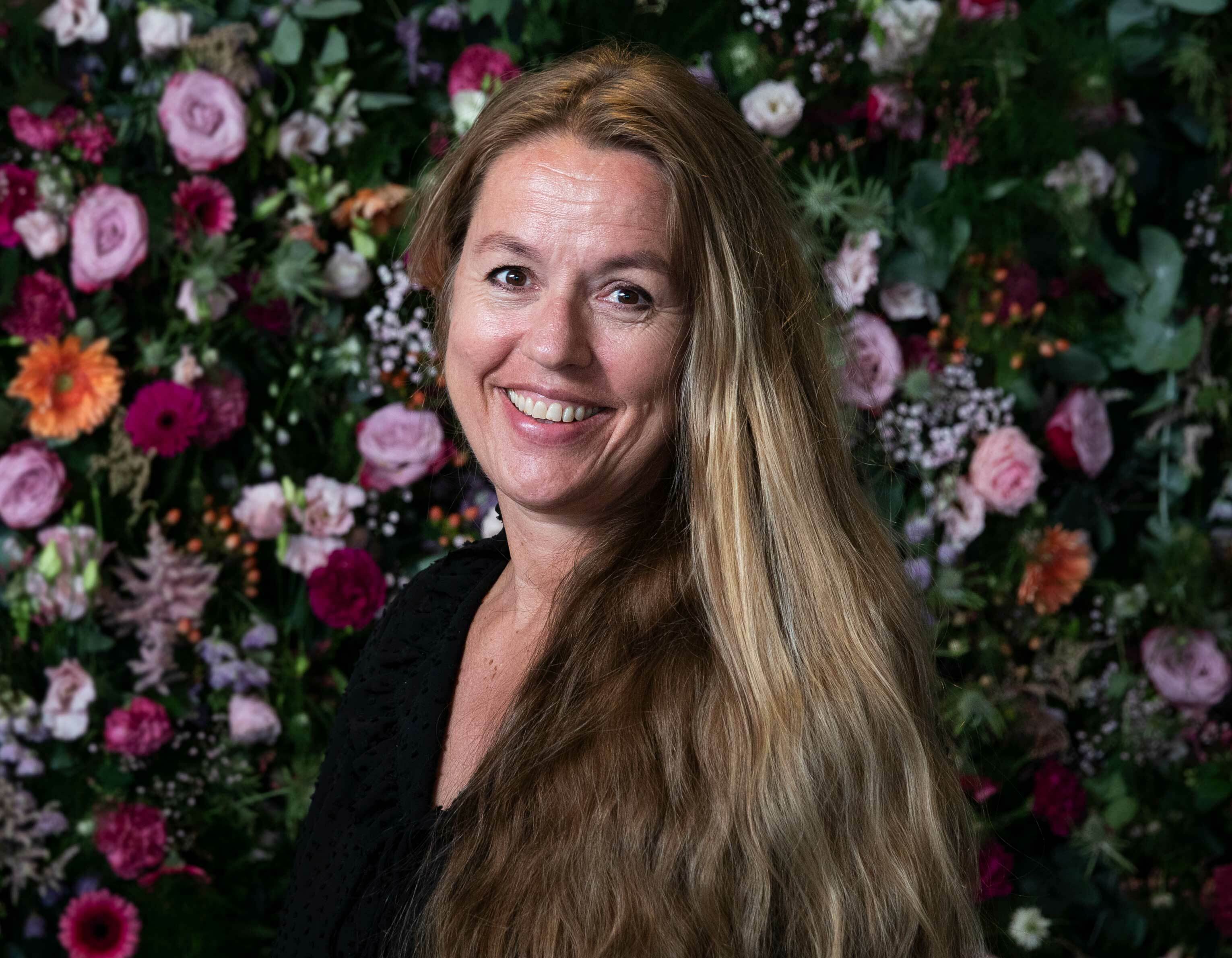 Foto: Portrett av Rita Bondeson Børke. Hun har langt, lyst hår og mørk jakke og smiler mot kamera. Bak henne er en vegg av blomster.