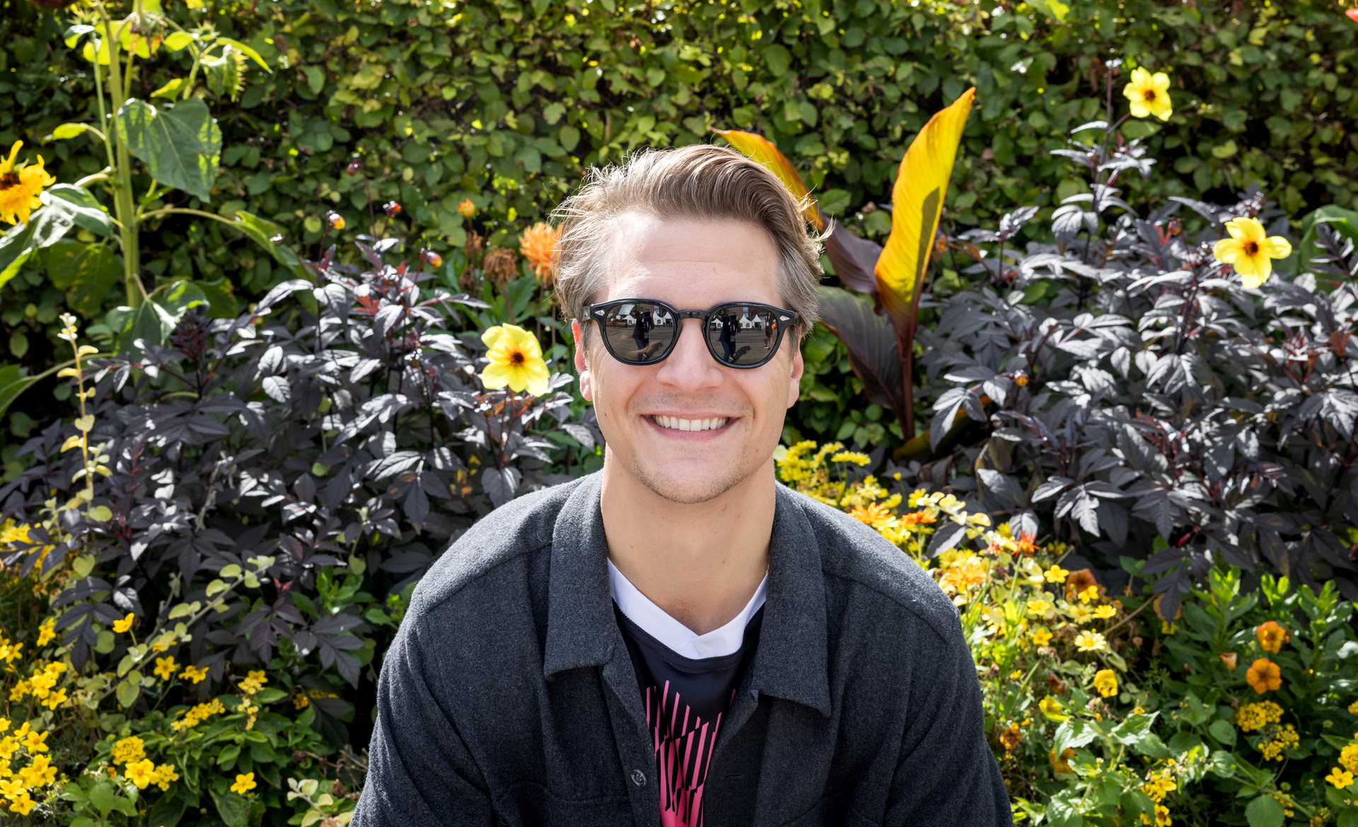 Foto: Blond mann i midten av 30-årene har på seg solbriller og smiler foran et blomsterbed med gule og oransje blomster.