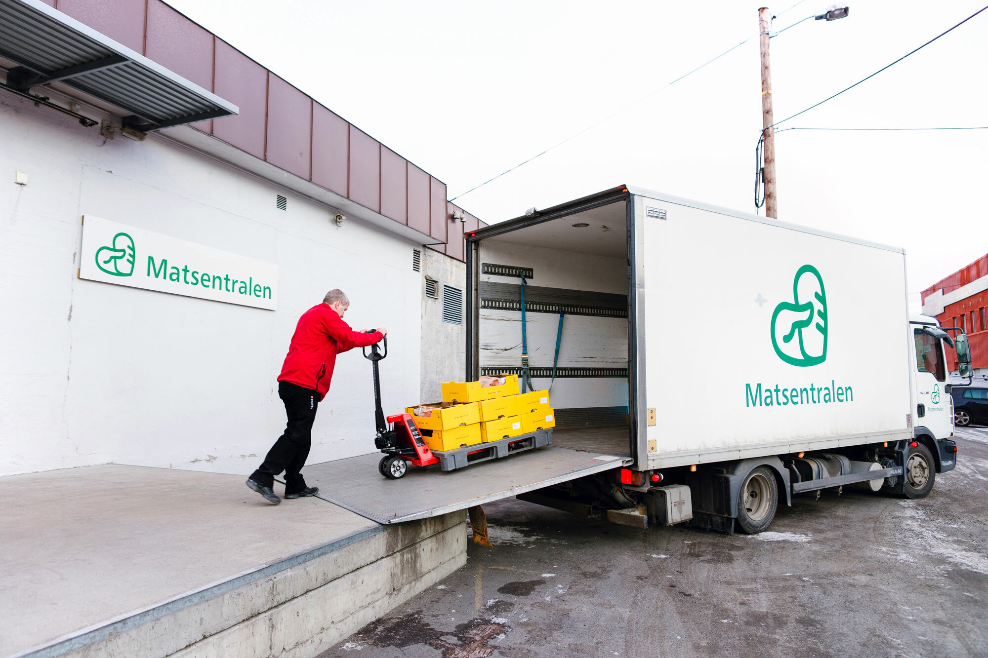 Foto: En mann i rød flisjakke triller en pall på en stor, hvit varebil. På siden av bilen, og på bygget det losses fra, står logoen til Matsentralen.