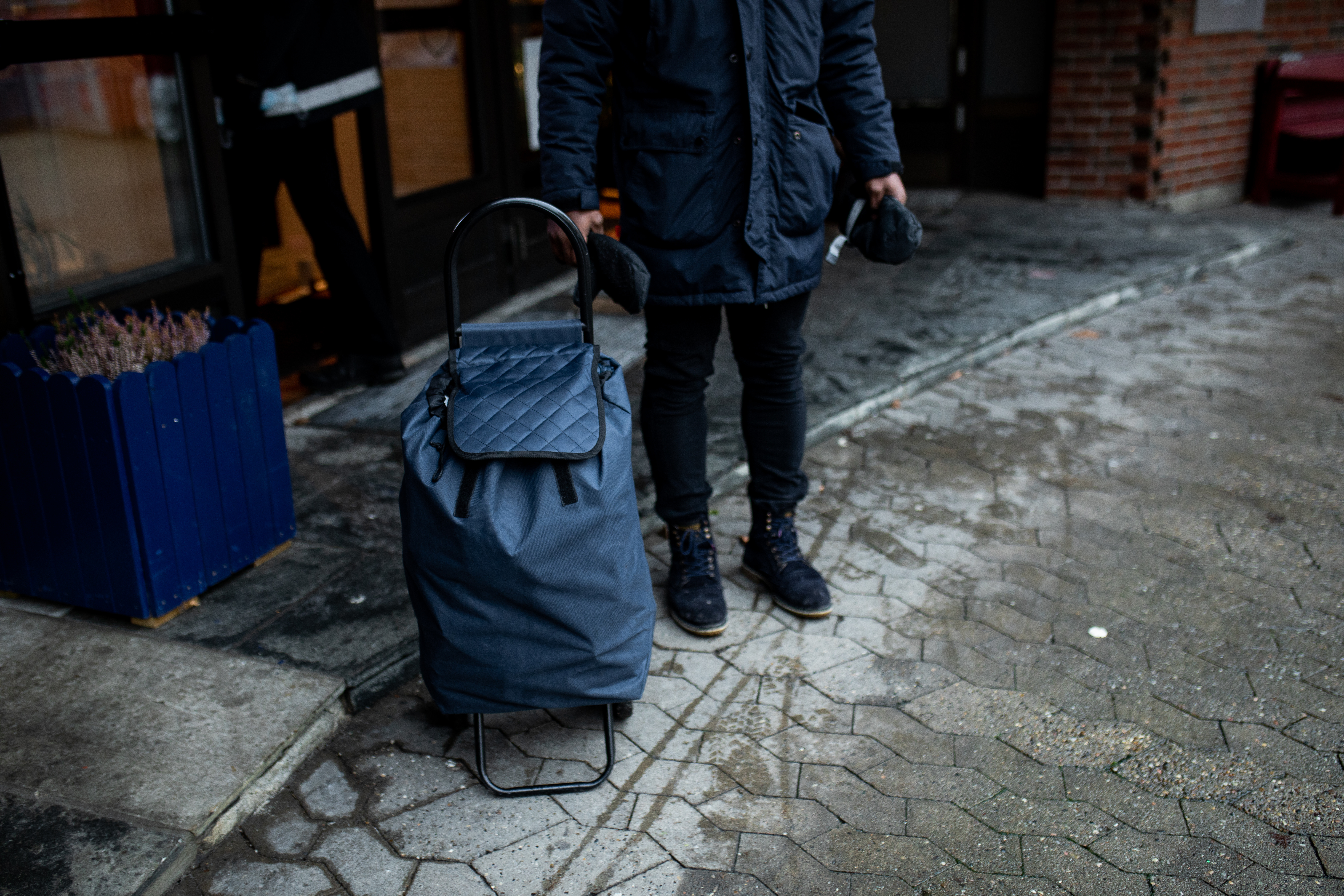 En person står foran en inngang i mørke klær og med en stor trillebag. Vi ser kun kroppen fra livet og ned.