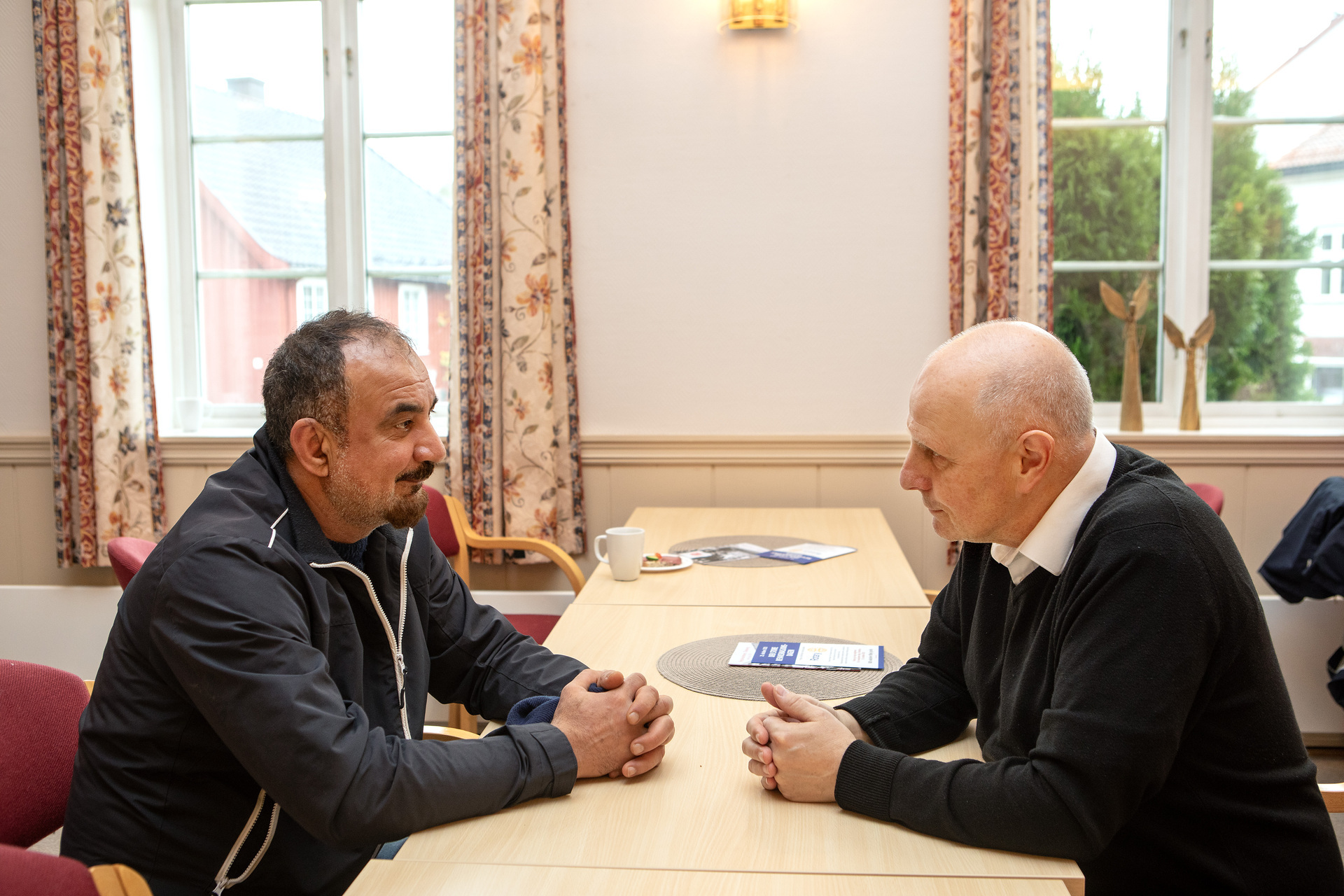 Foto: Bildet er tatt fra siden og viser Chris Pender som sitter ved et langbord og snakker med en mann. Begge har på seg svarte klær, de ser på hverandre mens de snakker.