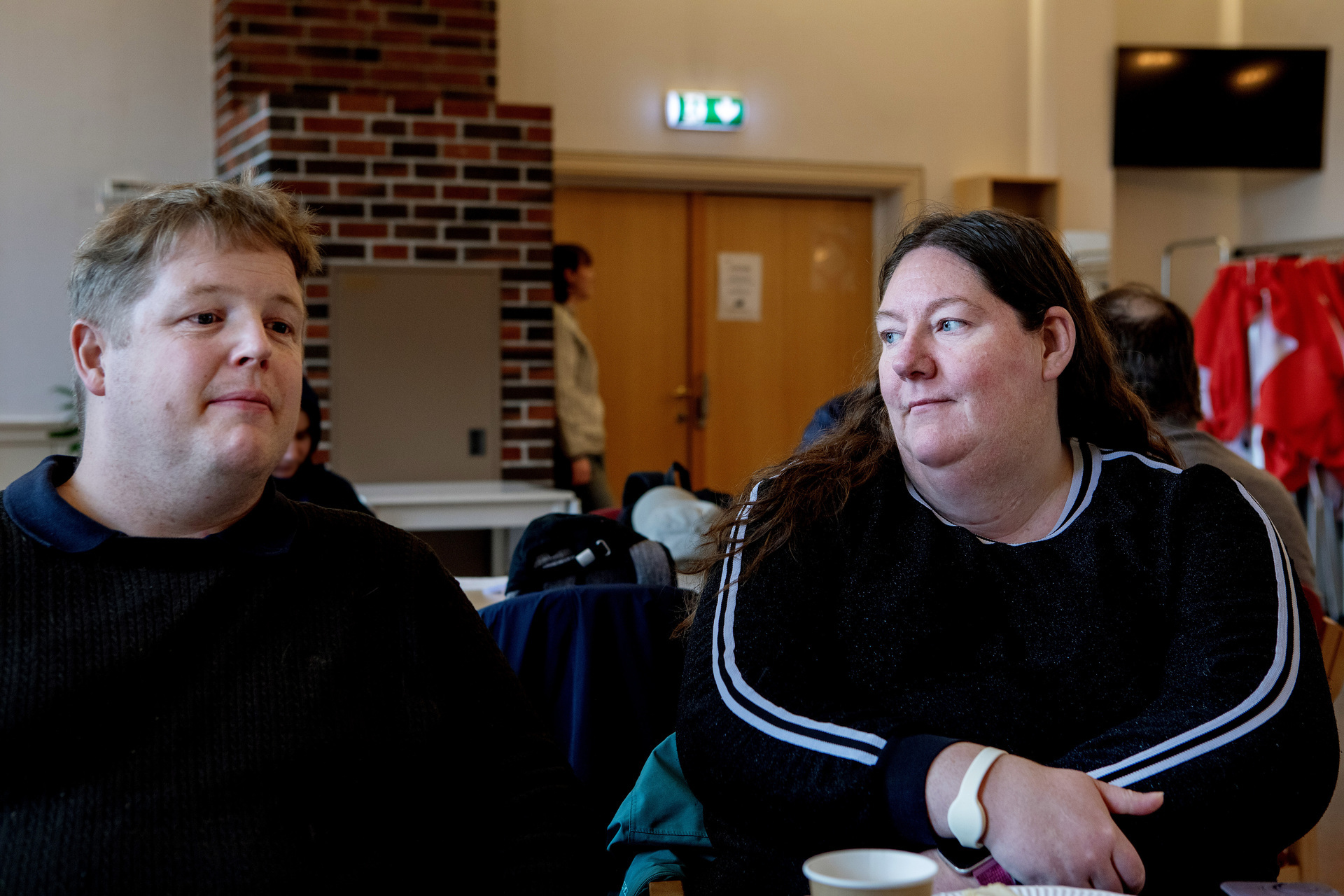 Foto: Jarle og Marita sitter ved et bord, Jarle har på seg svart genser og ser litt mot siden og Marita io svart genser med hvite striper sitter vendt mot ham. I bakgrunnen sees litt av lokalet til korpset, og døra inn til matutdelingen.