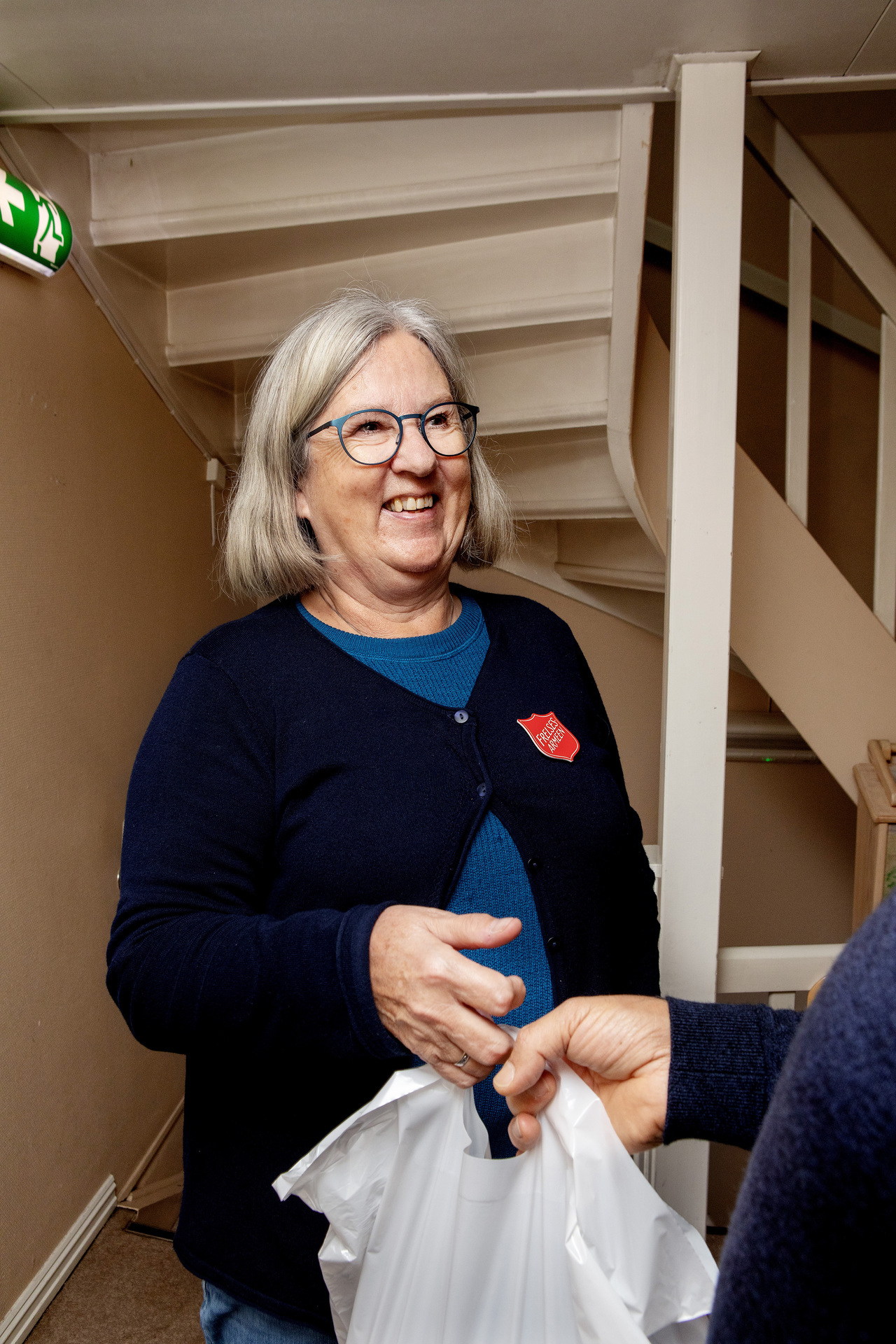 Foto: Gerd Britt deler ut en pose, hun smiler til personen som tar imot. Mottakeren er utenfor bildet, bare hånden hans synes. Gerd Britt har halvlangt, lyst hår og har på seg en svart strikkejakke med Frelsesarme-skjold.
