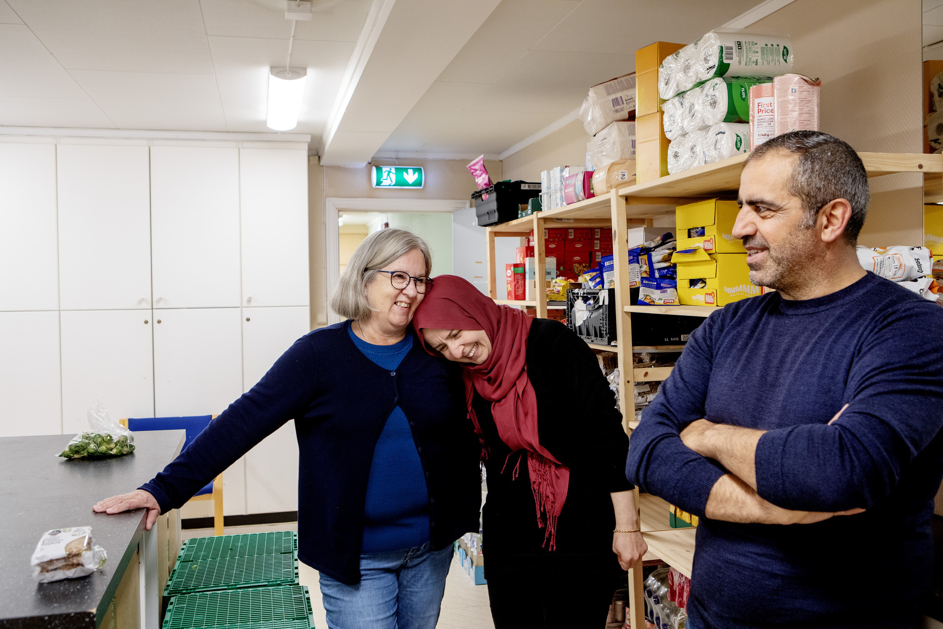 Foto: Buket i rød hijab og svart jakke står midt i bildet og lener seg mot skulderen til Gerd Britt. Fatih i blå genser står med armene i kors og ser mot dem og smiler. Bak dem sees hyller med mat og bordet der matposene nettopp har blitt pakket.