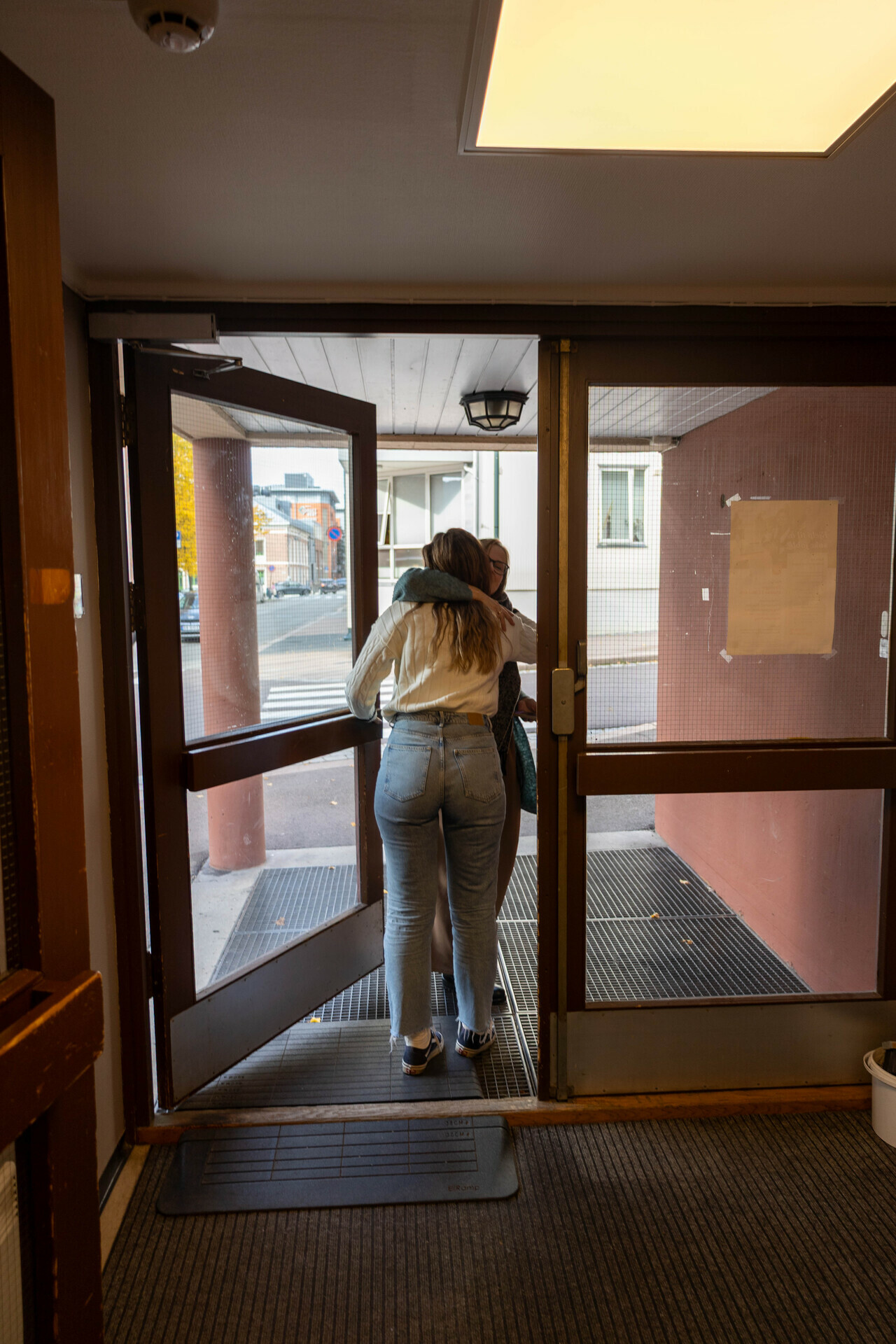 Foto: Dhurata gir Inese en god klem i døra på vei ut fra Frelsesarmeens lokaler. En gate og et hvitt hus sees i bakgrunnen. Dhurata står med ryggen til kamera, Inese sees så vidt bak henne.