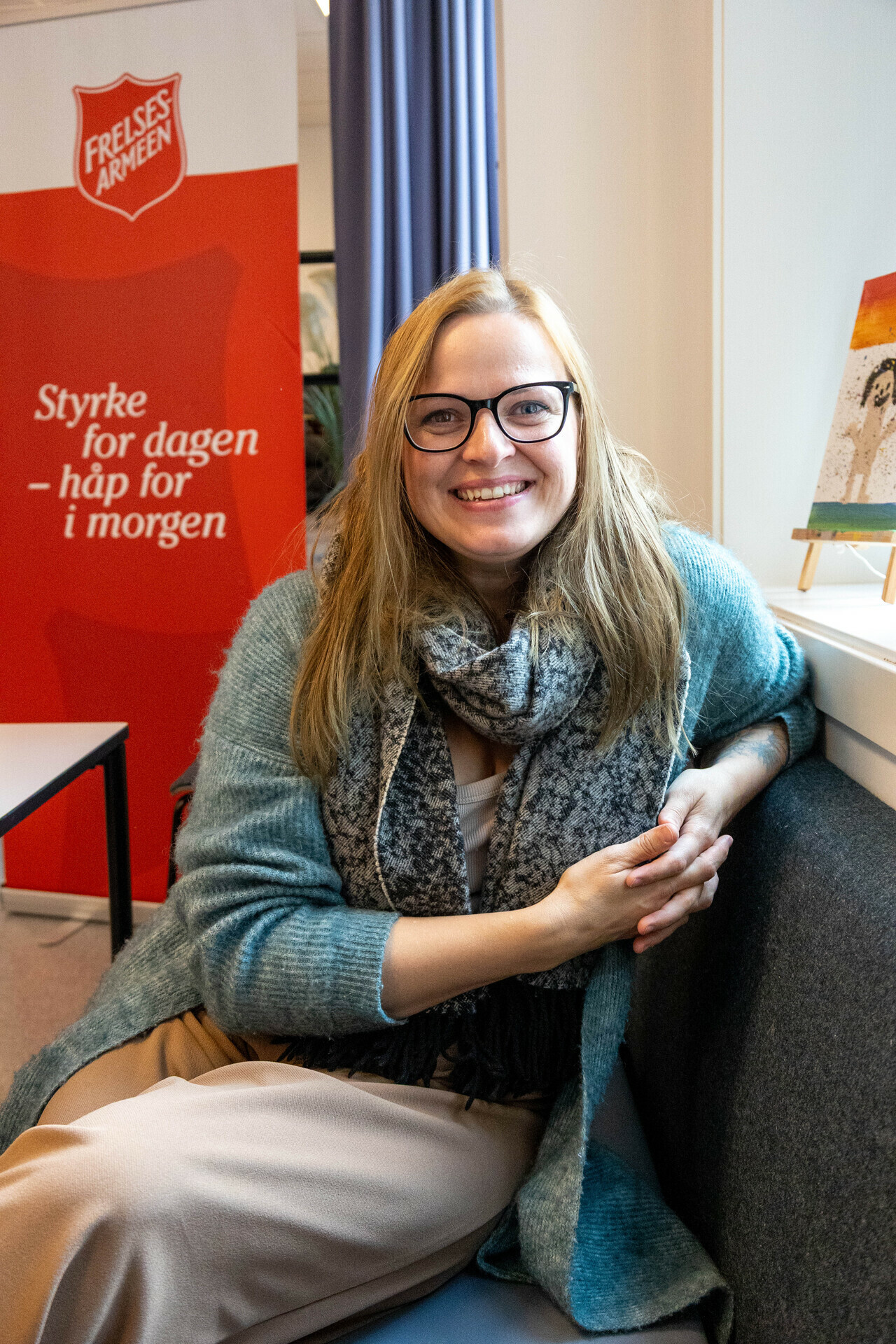 Foto: Inese sitter I en sofa vendt mot kamera og smiler. Bak henne sees et stort banner med Frelsesarme-logo og teksten Styrke for dagen, håp for i morgen.