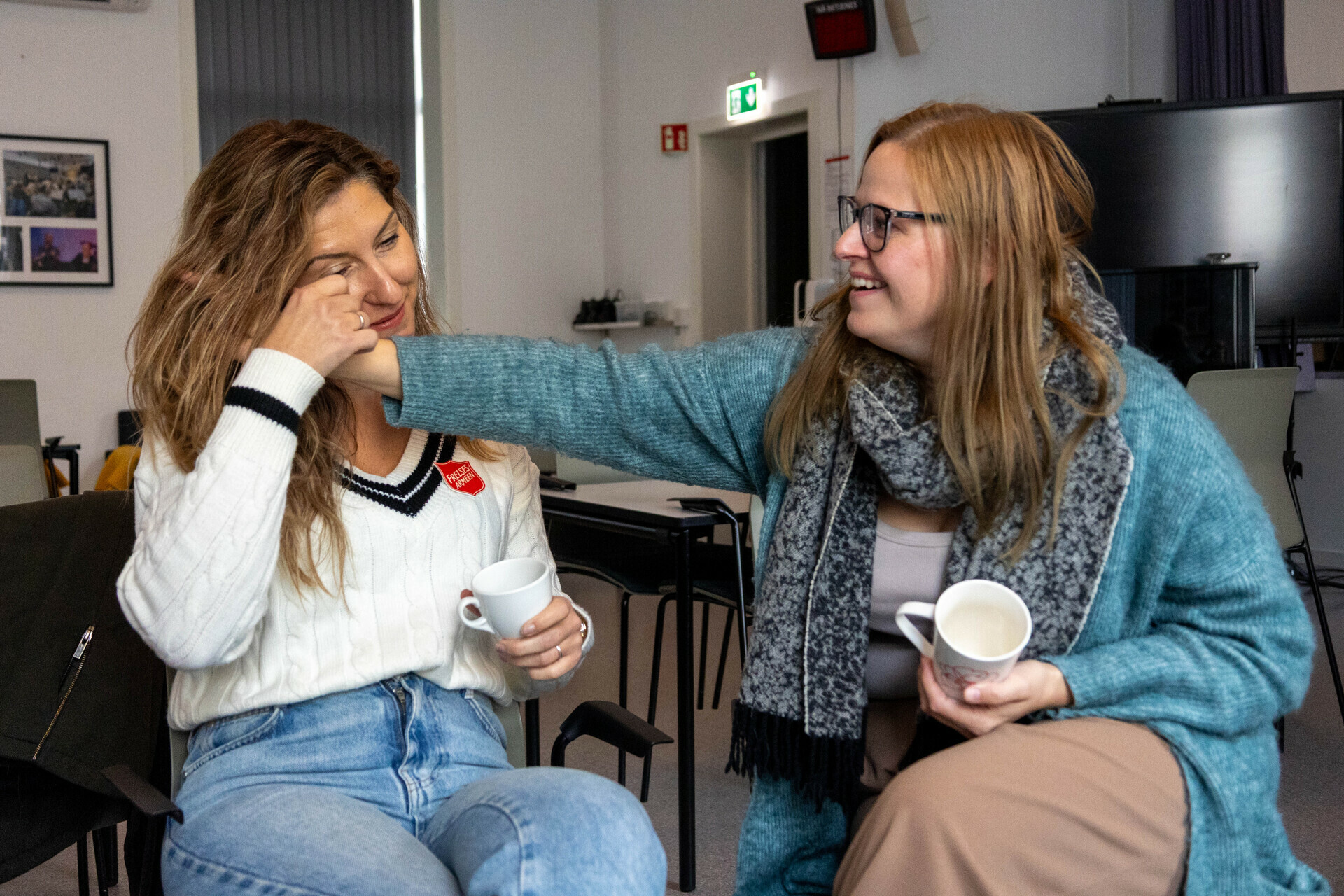 Foto: Dhurata sitter og holder en hånd opp mot øyet for å tørke en tåre. Inese ser på henne og smiler, og holder en hånd mot ansiktet hennes,