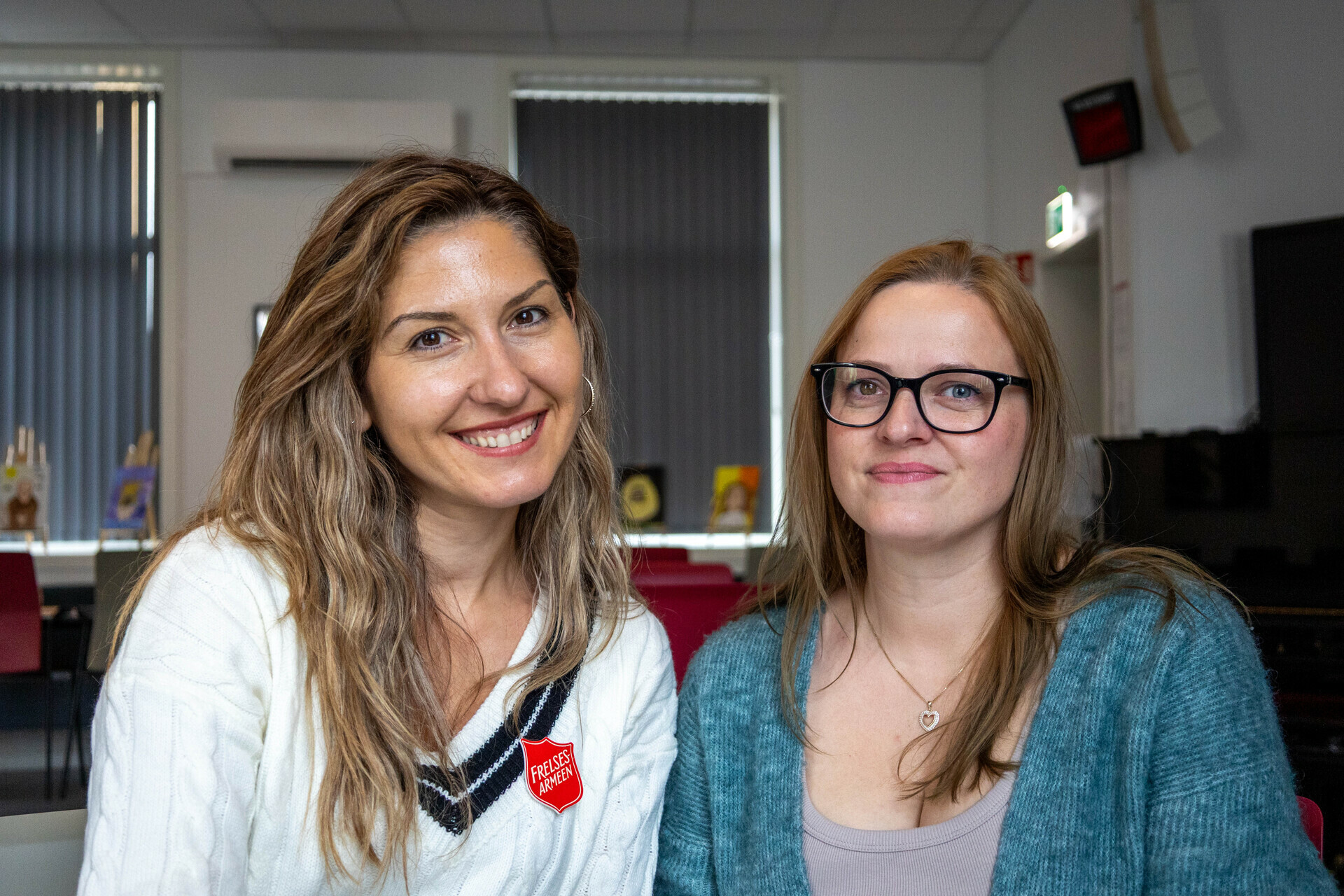 Foto: Dhurata Perdja Inese Kungurova sitter ved siden av hverandre og smiler mot kamera. Begge har langt hår, Dhurata har på seg hvit genser med Frelsesarme-skjold og Inese har på seg en grønn strikkejakke.