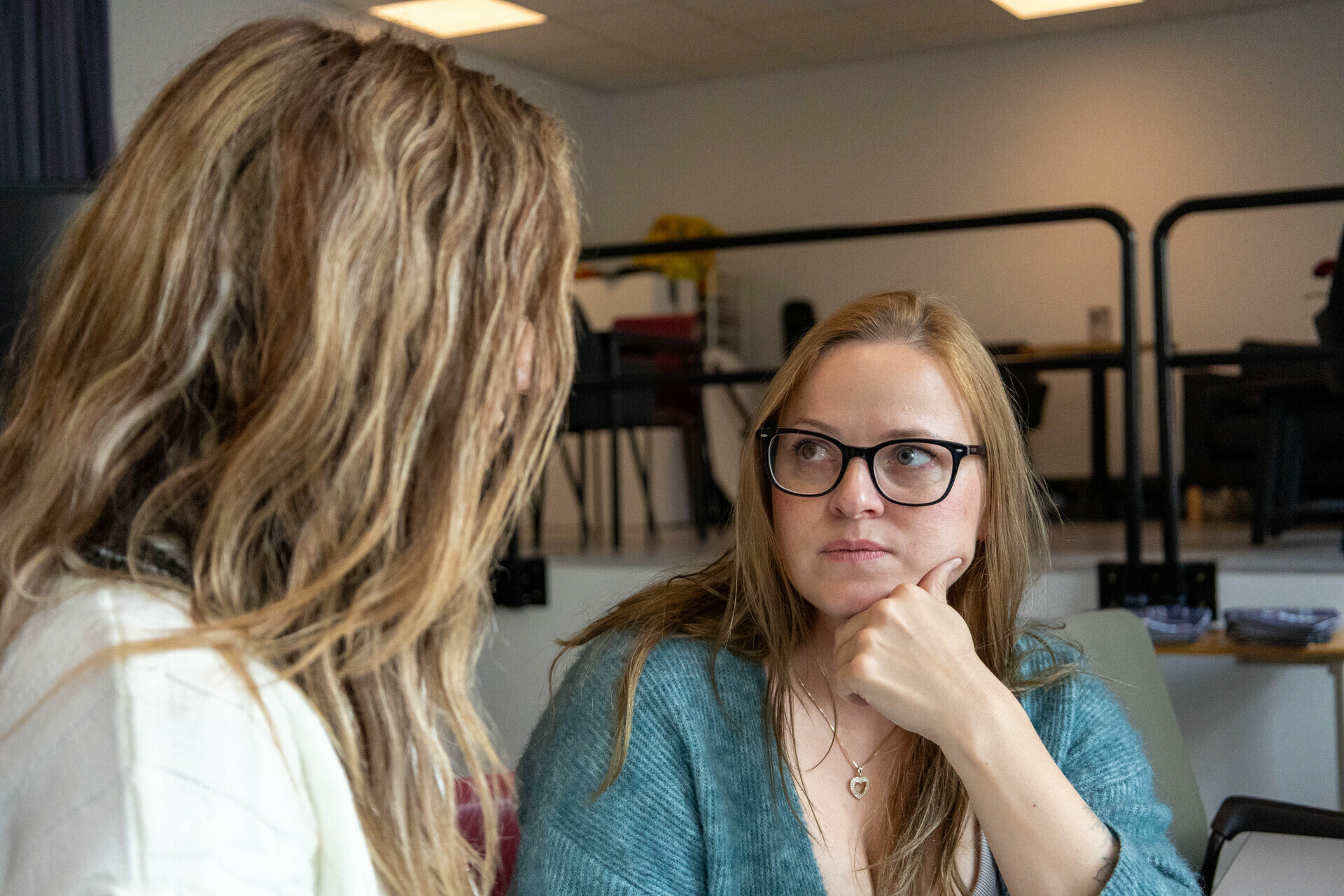 Foto: Dhurata sitter nærmest kamera med rygge mot, bare håret og litt av genseren hennes syntes. Inese sitter og ser mot Dhurata med et alvorlig ansiktsuttrykk.