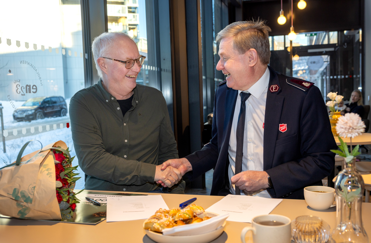Foto: To menn i 60-årene, en med briller og grønn skjorte, den andre i uniform fra Frelsesarmeen sitter ved et bord i en kafe. På vinduet står logoen til Kafé En23.