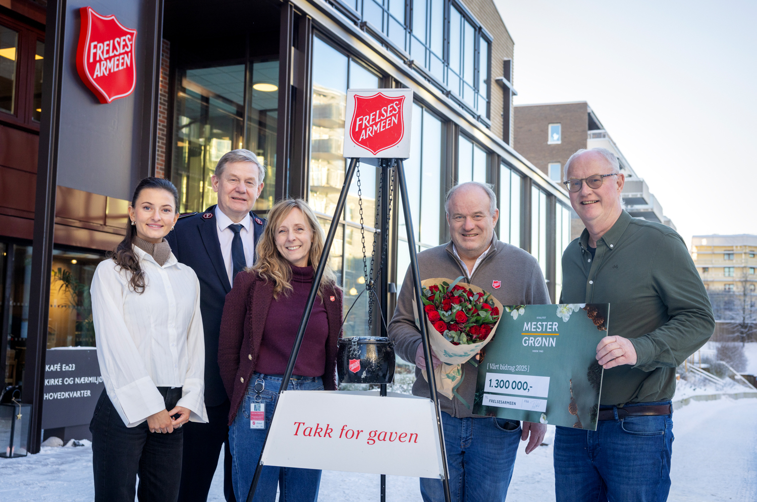 Foto: Fem personer, to kvinner og tre menn, står på rekke ved et stativ med en julegryta fra Frelsesarmeen. En av mennene har på seg uniform og de andre to holder en sjekk og en blomsterbukett.