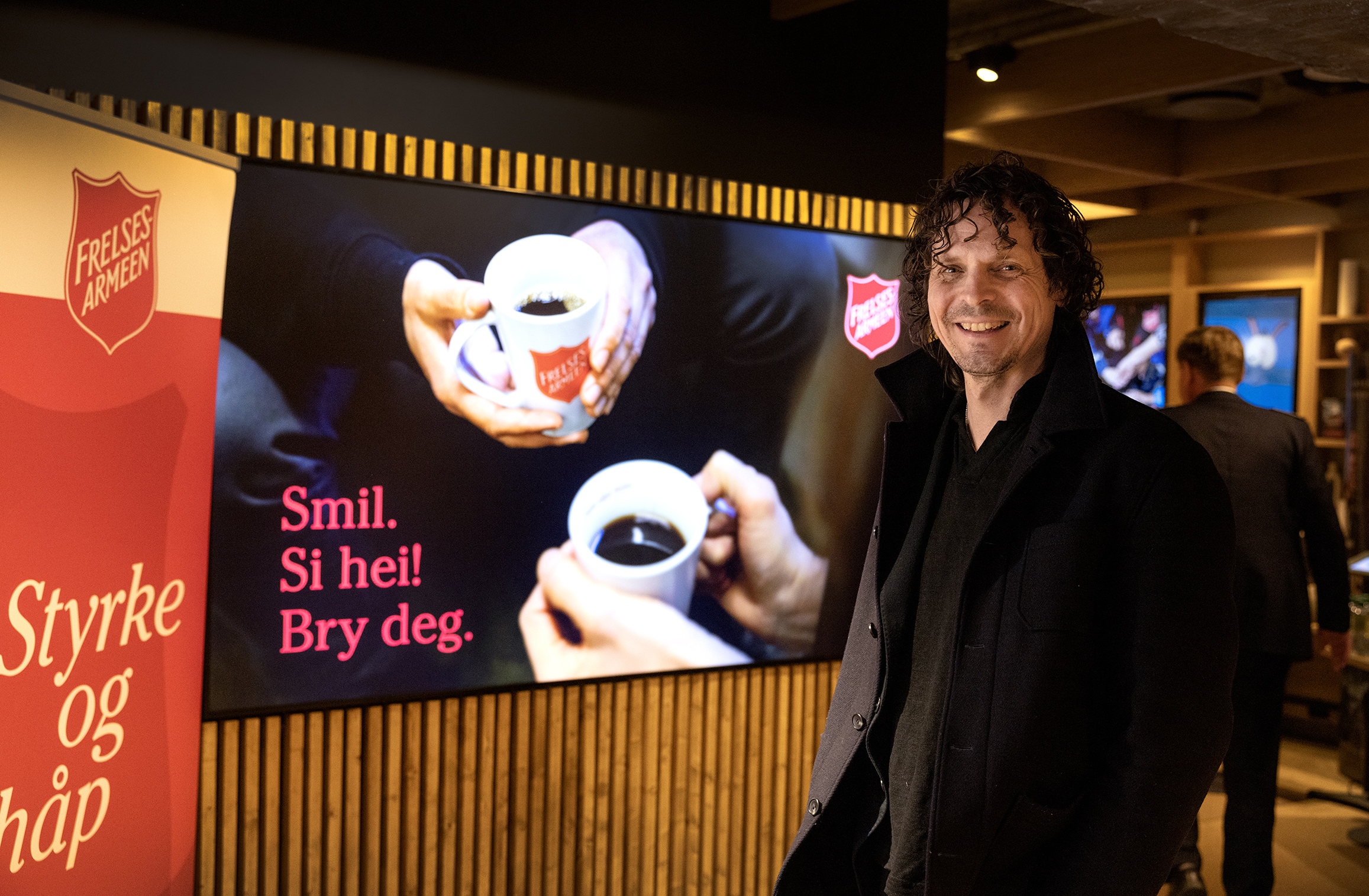 Mann står i kinoens foaje. På skjermen bak er et bilde med hender som holder kaffekopper. Det står skrevet: Smil. SI hei! Bry deg.