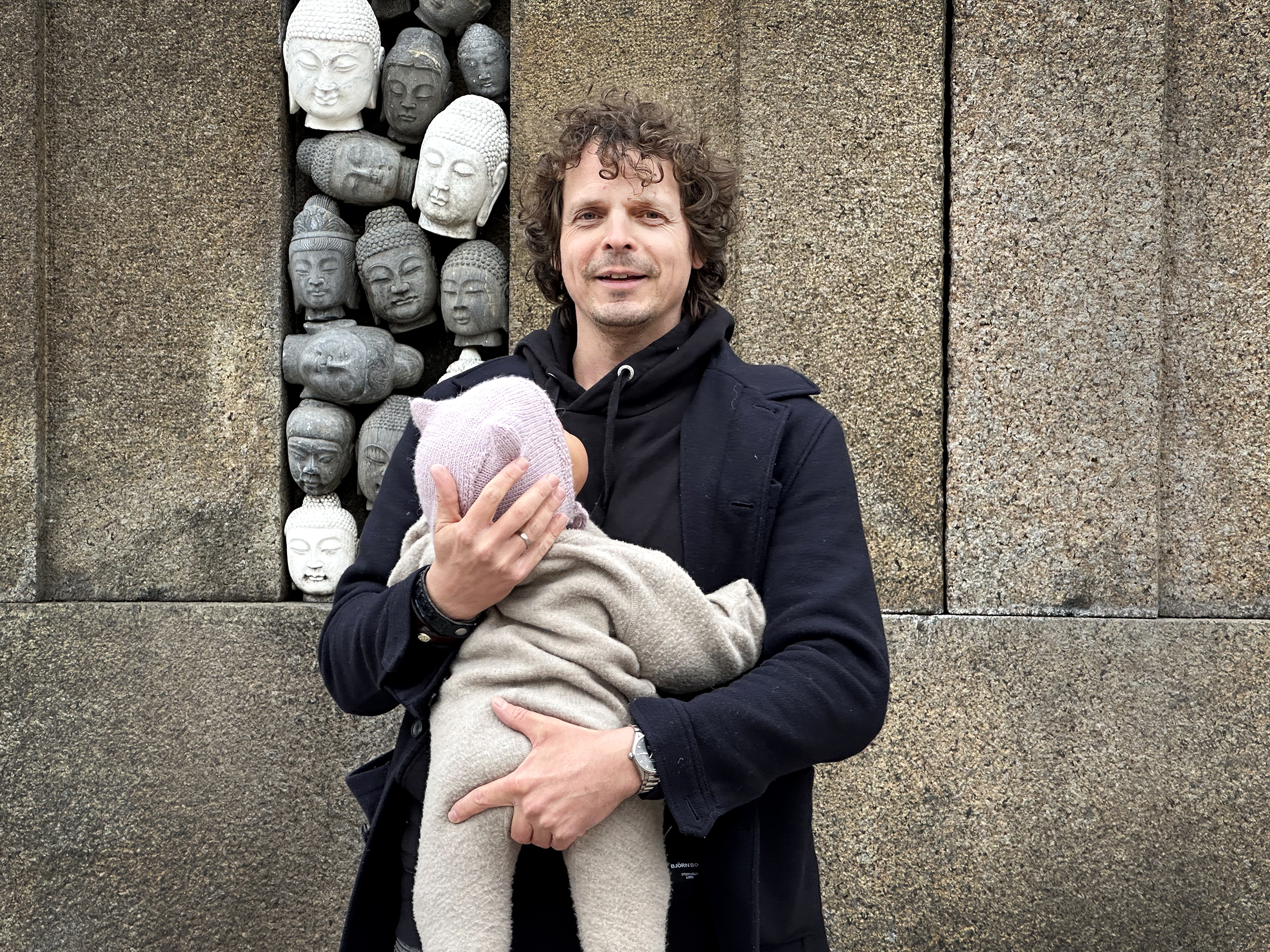 Mann holder baby utendørs. Han ser rett i kamera, ungen ser på ham. I bakgrunnen mange Buddha-hoder stablet i en stor steinblokk i en park.