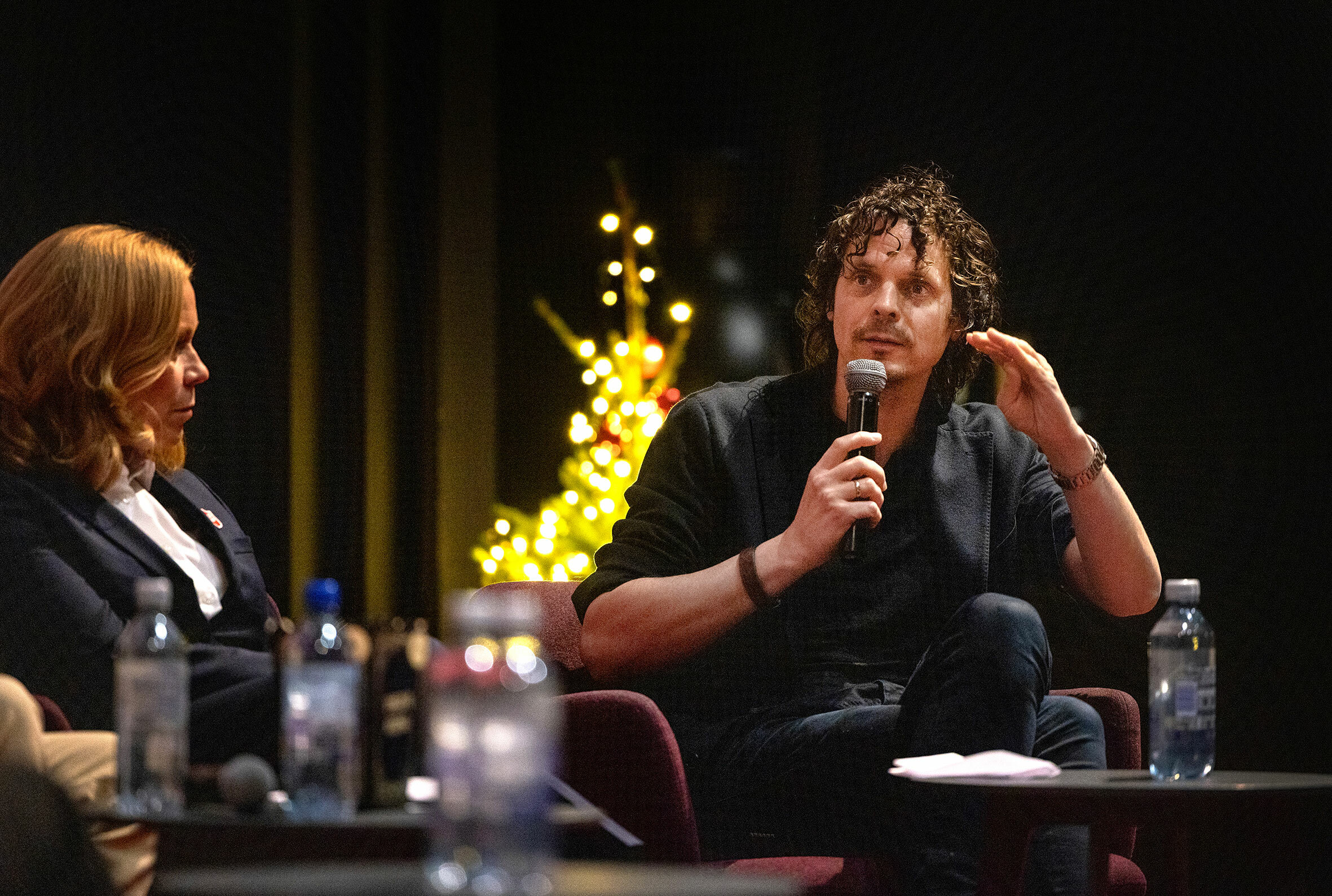 Mann snakker i mikrofon. I bakgrunnen står et lite juletre med tenkte lys. Dame i stolen ved siden av lytter.