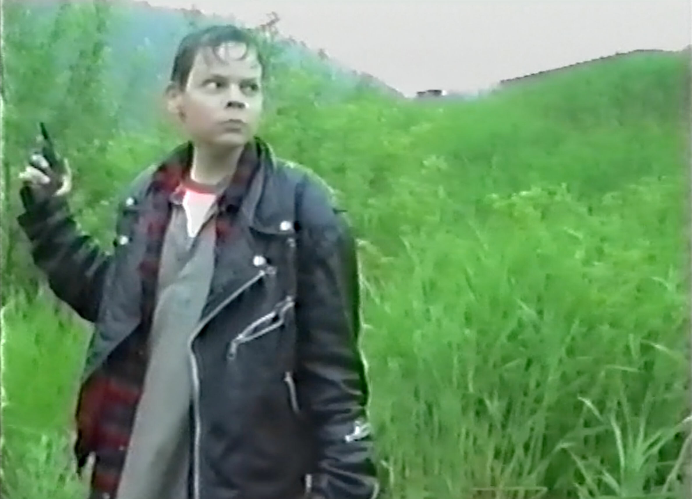 Ung gutt i skinnjakke. Står i høyt, grønt gress med en pistol i hånda.