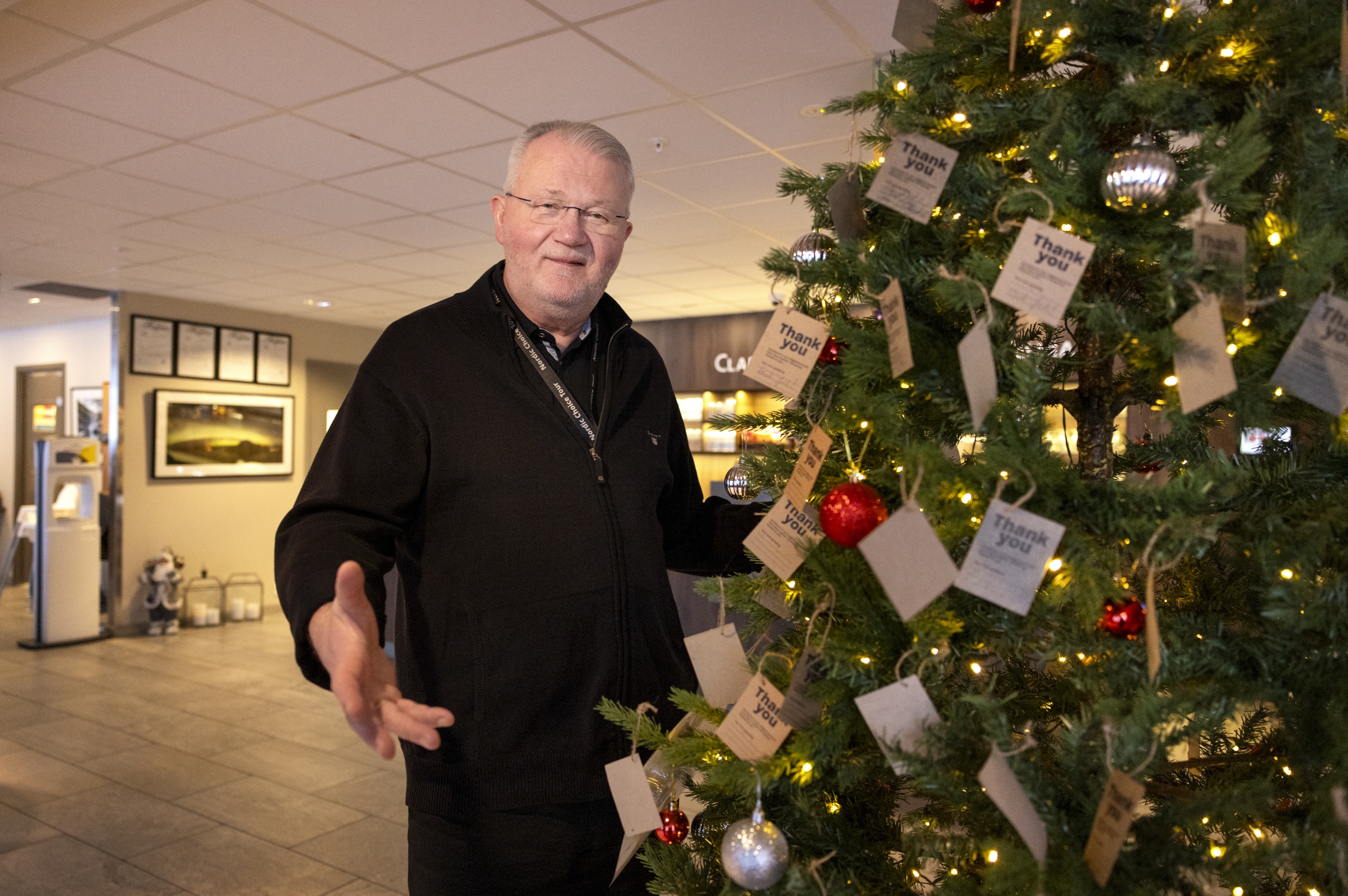Mann står med hender utstrakt ved et juletre. Her henger det mange julepynt og lapper.