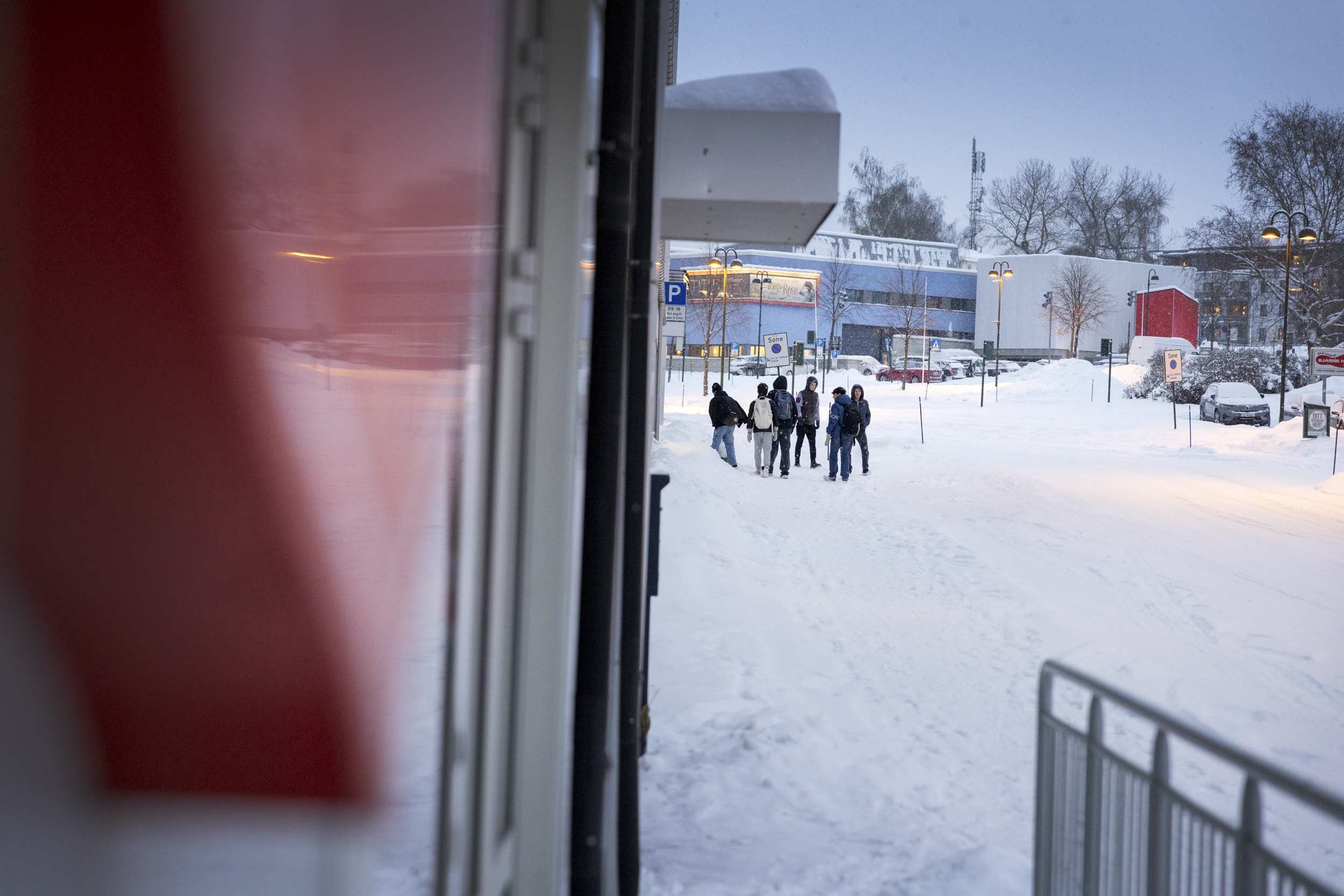 Dør står åpen, ut ser man ungdommer som går oppover i en gate dekket av snø. På andre sida av for dem står noen få biler parkert på en tomt.