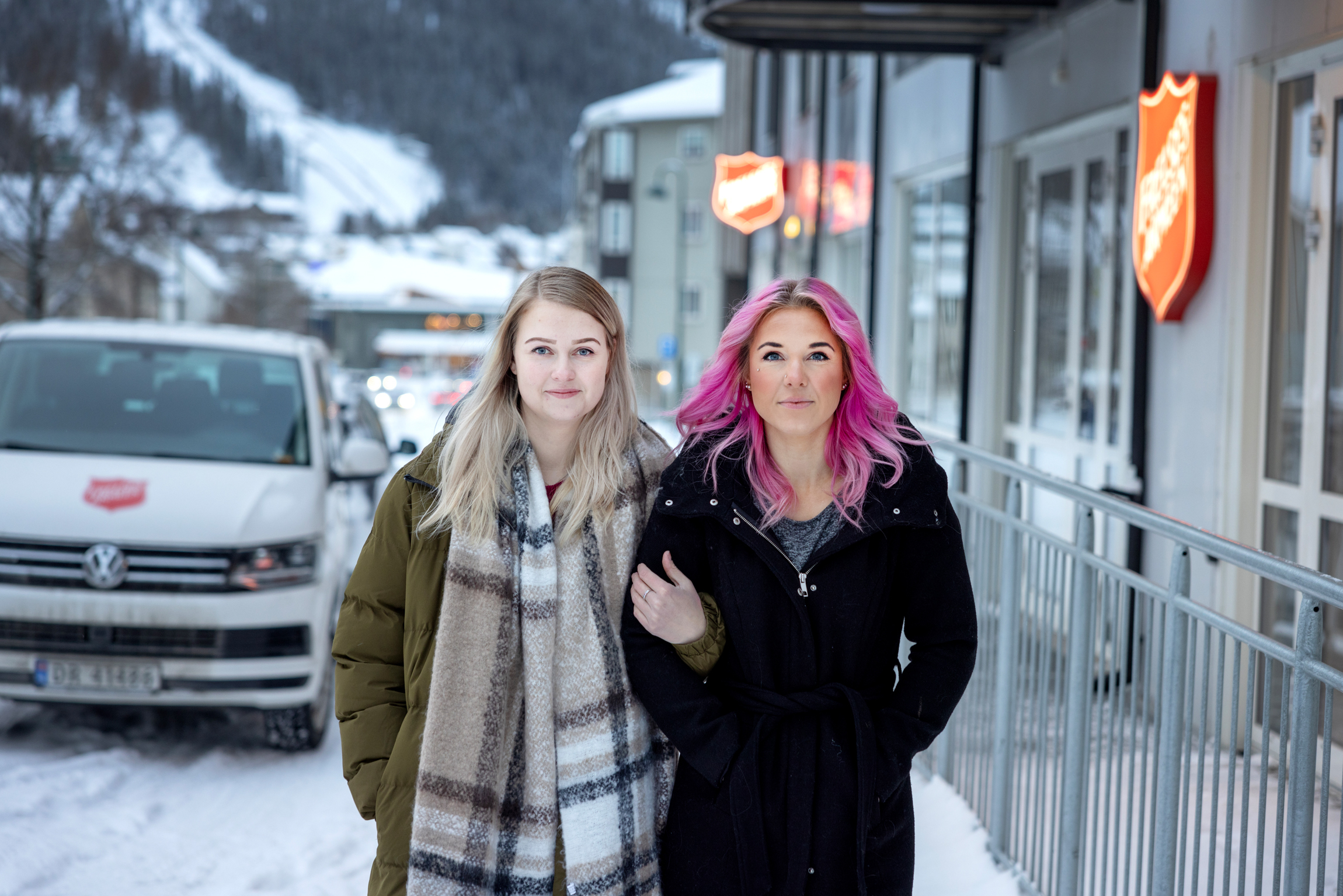 To kvinner står arm i arm utendørs utenfor Frelsesarmeens lokaler. Det er mye snø og en hvit minibus med Frelsesarmeens logo på.