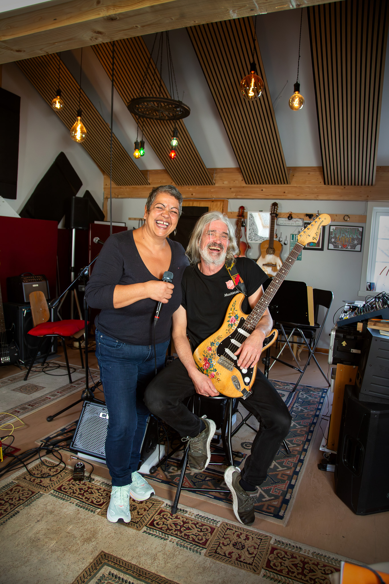 Foto: Kvinne og mann smiler og ler. De er i et rom fylt med instrumenter. Mannen holder i en el-gitar med blomsterdekorasjon på.