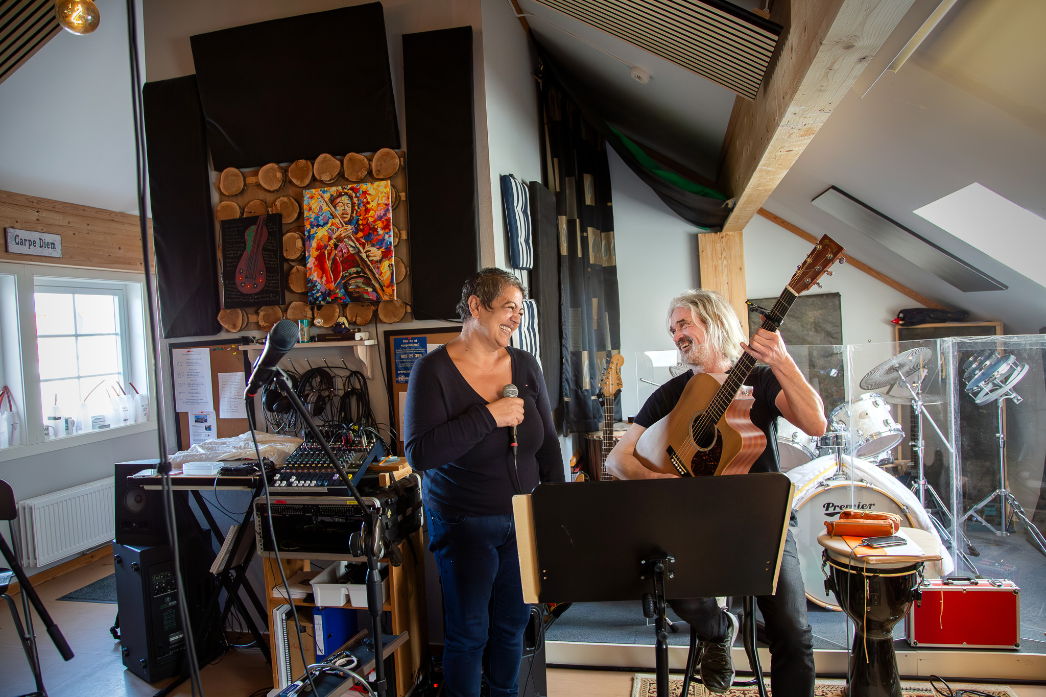 Kvinne og mann står inne i et rom fylt med instrumenter. Kvinnen synger og mannen spiller gitar