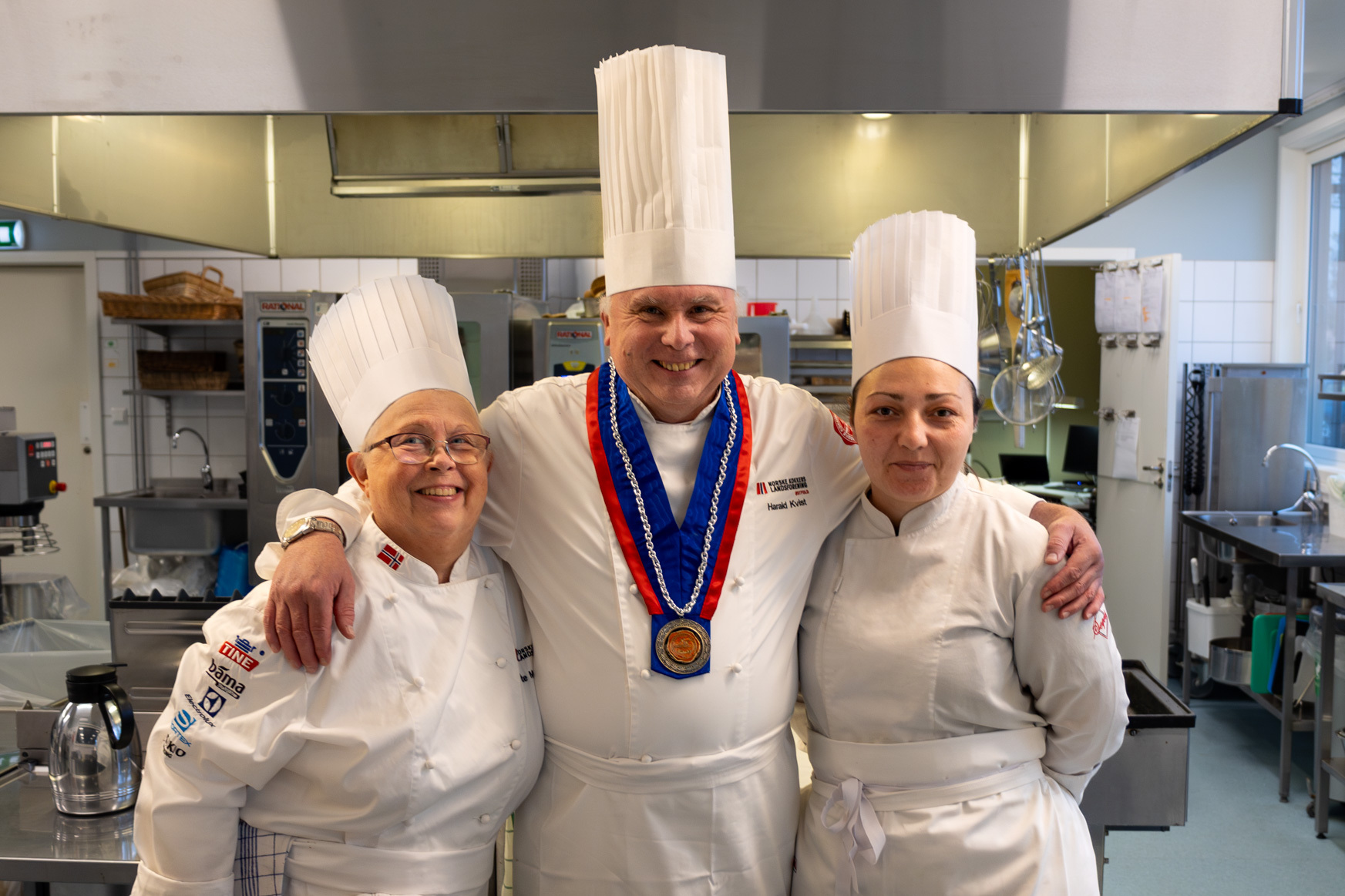 Foto: Tre kokker i hvite drakter med hatter, to kvinner og en mann. Han står i midten og holder armen rundt begge. Alle smiler.
