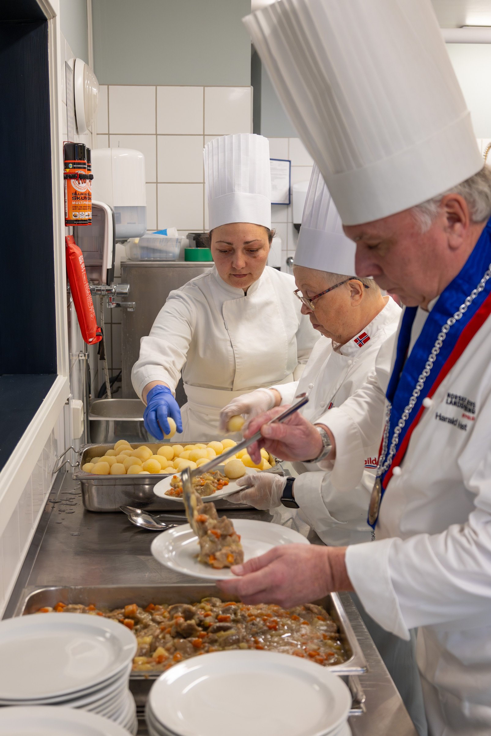 Foto: Tre kokker, en mann og to kvinner, står på rekke og legger mat på en tallerken.