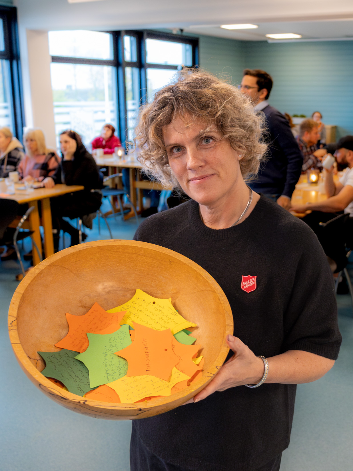 Foto: Kvinne med krøllet hår, svart genser og logo fra Frelsesarmeen holder frem en skål. Oppi ligger det ark med mange farger.