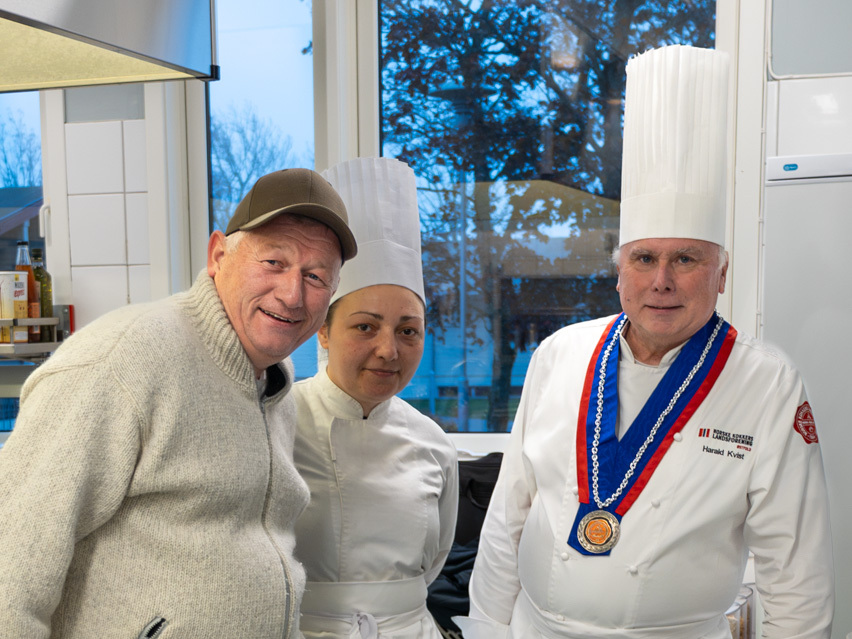 Foto: Mann med caps og ullgenser smiler fra kjøkkenet. Han står sammen med to av kokkene, en kvinne og en mann.