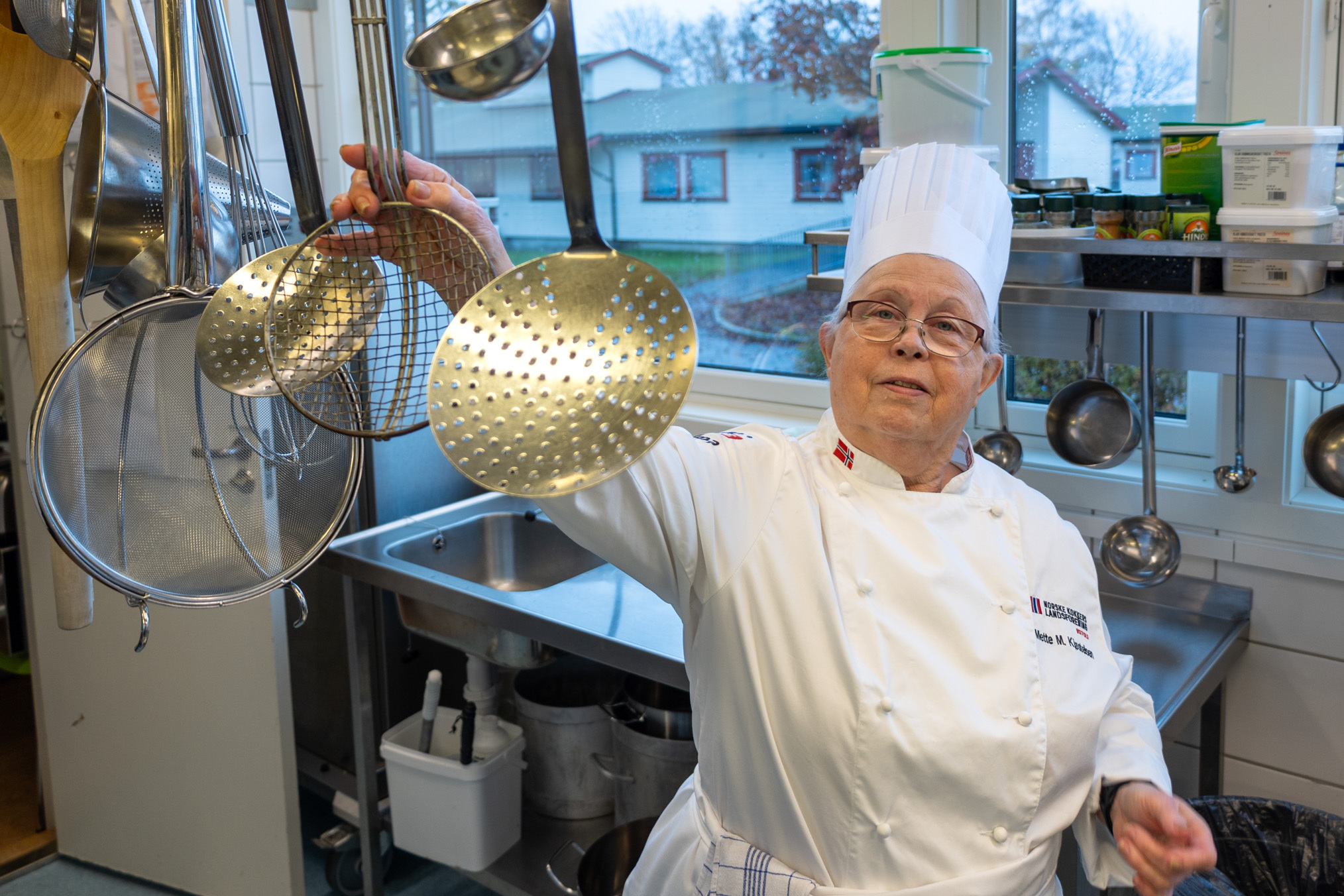 Foto: Kokk i 70-årene med hvit drakt og forkle strekker seg etter en øse og flere andre redskaper som henger på en rekke midt i industrikjøkkenet.