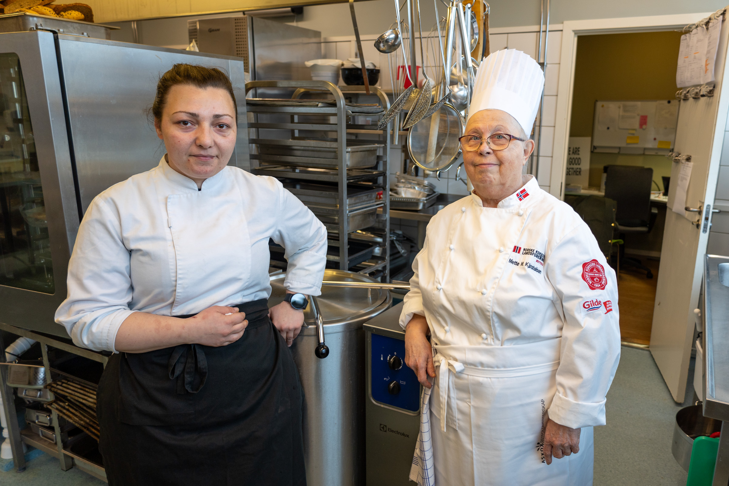 Foto: To kvinelige kokker står ved siden av hverandre. Begge ser i kamera. Den ene er i ferd med å ta av seg forkleet.