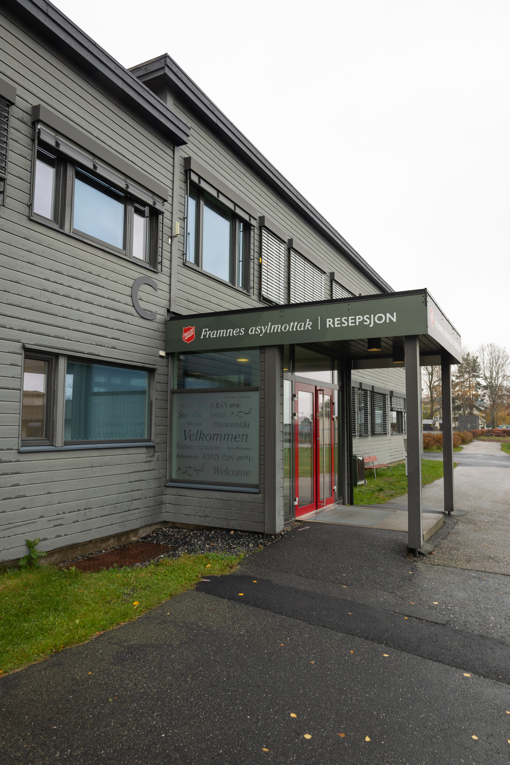 Foto: Utvendig bilde tatt av grått bygg med flatt tak. Vinduene har røde karmer og over et inngangsparti står det "FRamnes asylmottak – resepsjon" ved siden av en logo fra Frelsesarmeen.