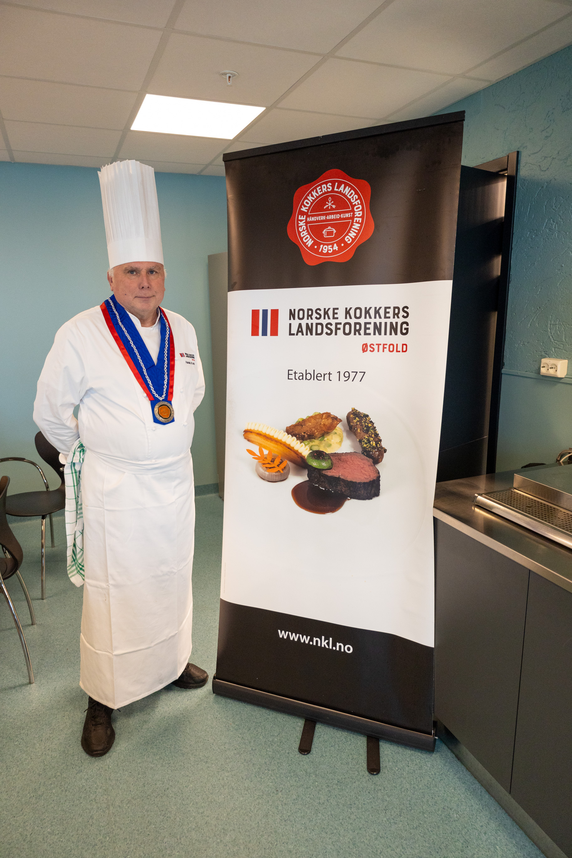 Foto: Kokk som står oppstilt ved siden av en reklameplakat med en pent dandert matrett på og et par logoer.