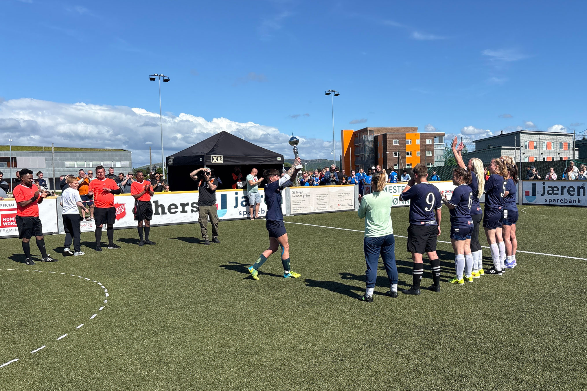 Foto: Spillerne på et kvinnelag står oppstilt på en bane med ryggen til kamera. Folk står og heier og klapper. En fotograf tar bilder mens ene spilleren holder oppe enstor pokal. Det er knall blå og nesten skyfri himmel.