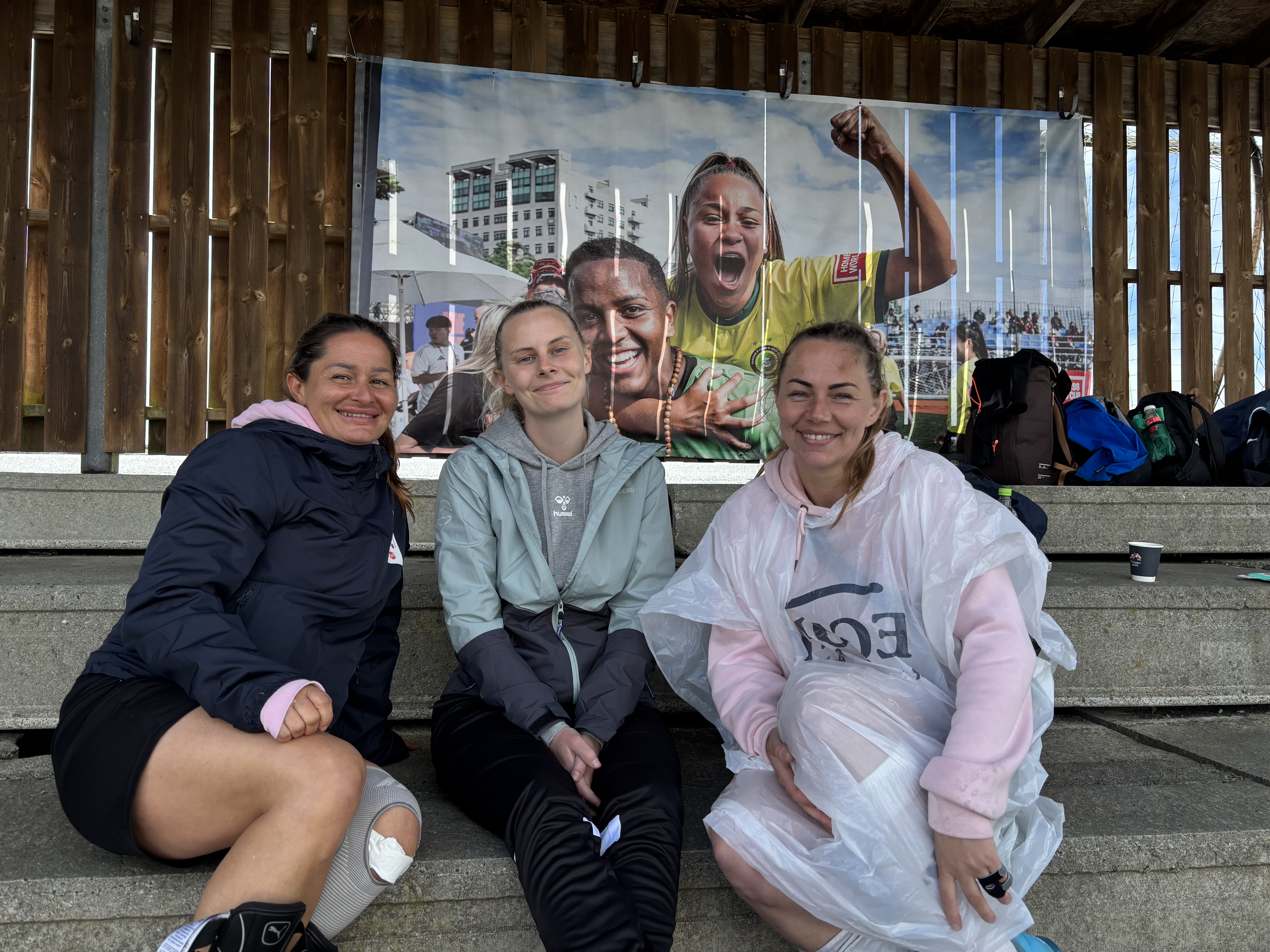 Foto: Tre unge, smilende kvinner i regntøy på en tribune. På bakvegge i tre henger et bilde fra et tidligere Homeless World Cup, eller VM i gatefotball.