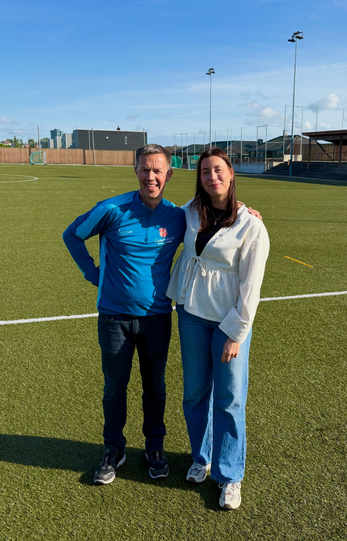 Foto: På en fotballbane står en mann og kvinne. Han har på seg blå treningsjakke og hun en hvit bluse. Begge har joggesko og jeans. De er tidlig i 30-årene.