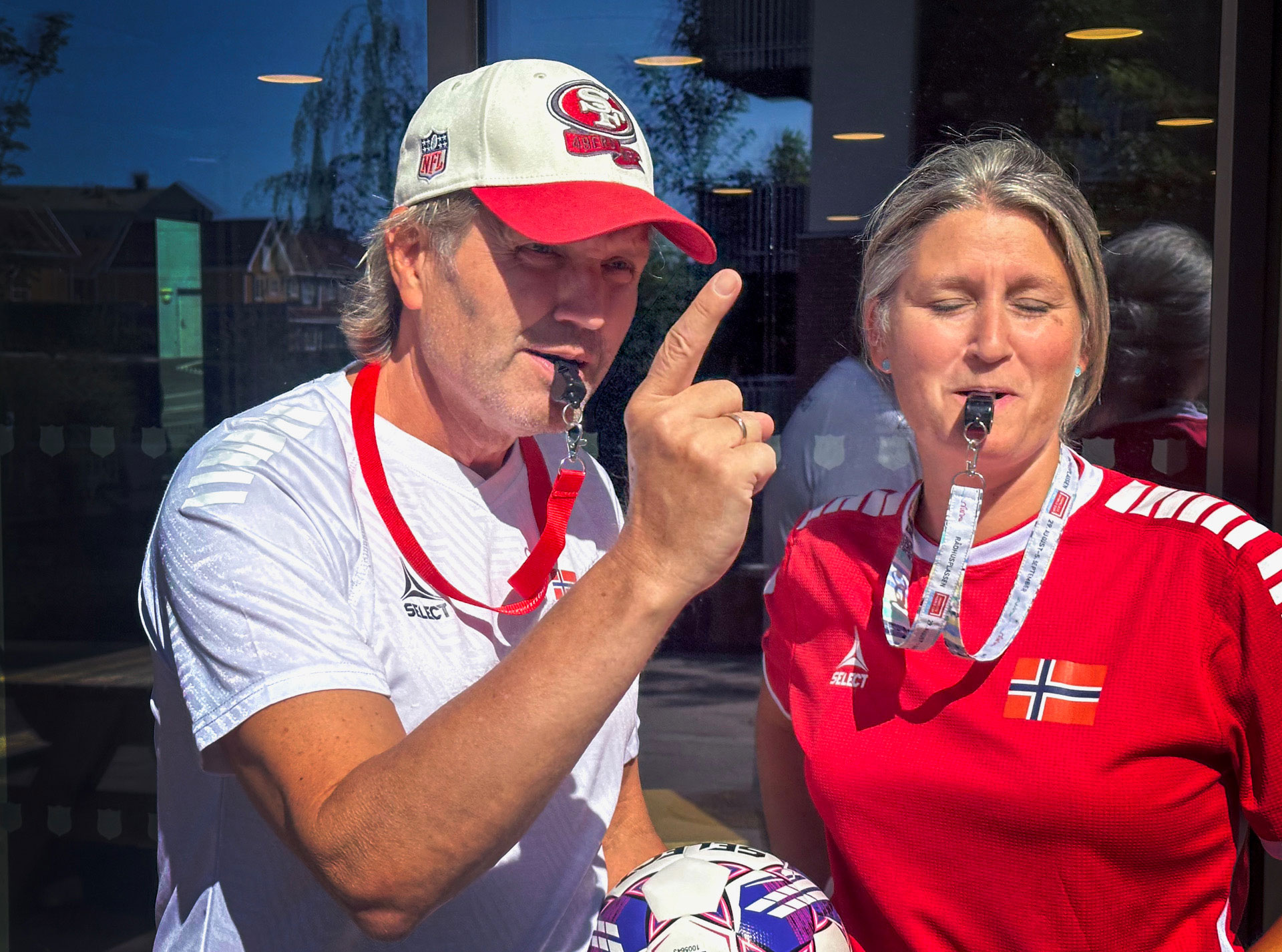 Foto: Mann i hvit drakt og kvinne i rød drakt. Han holder i en fotball og begge blåser i hver sin fløyte. De står ute, foran et vindu i sola. Han har på seg hvit caps med rød skygge.