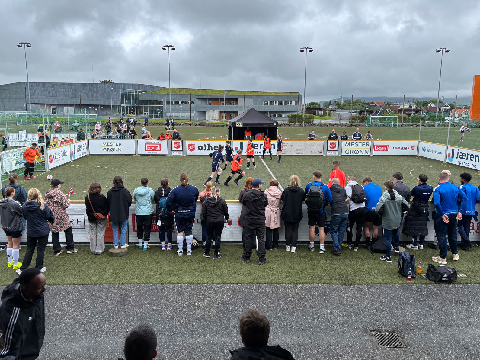 Foto: Det er mørke skyer og regne over en fotballbane. mange folk står og ser på kampen i regntøy.