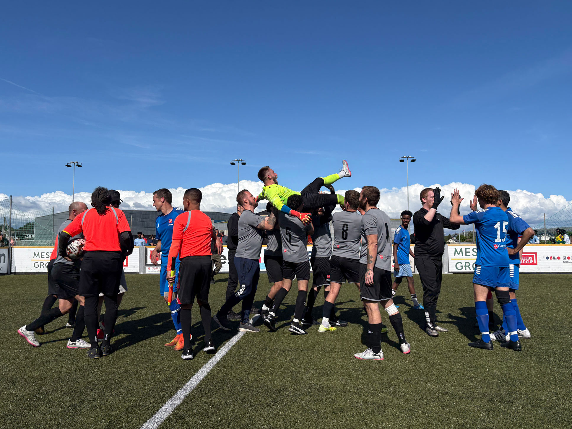 Foto: Mange fotballspillere samlet i midten av banen i litt ulike drakter. En mann i gult løftes i været av gjengen i grå drakter.