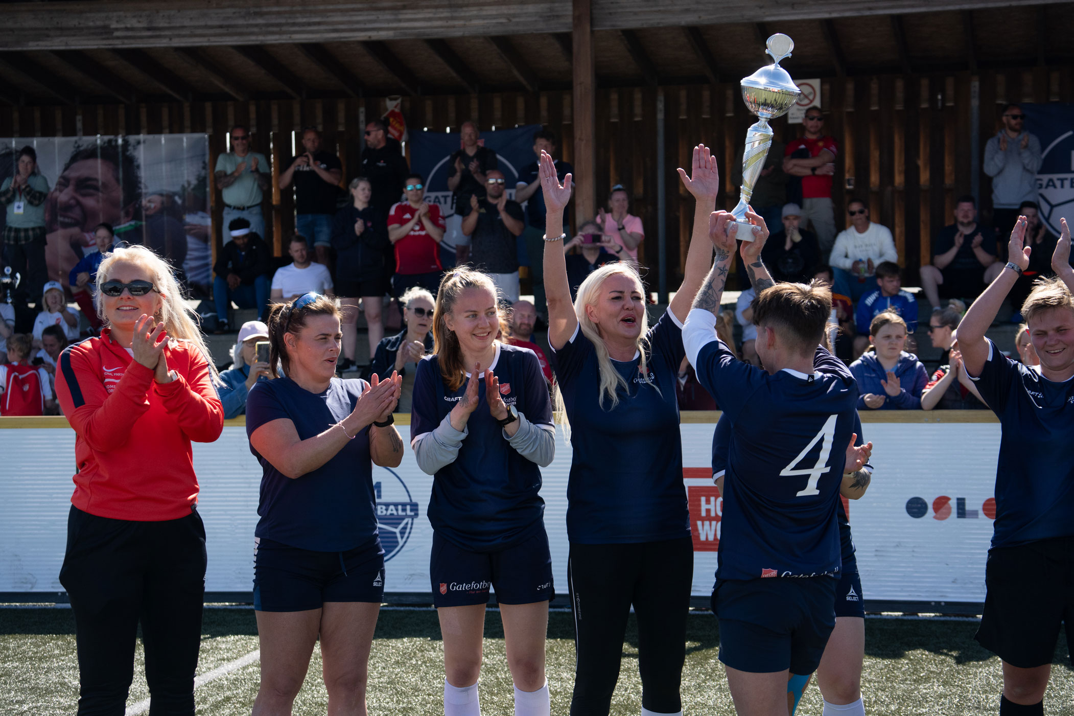 Foto: Syv kvinner i fotballdrakter står på rekke inne på en gressbane med publikum på tribunen bak. De står med ansiktene mot fotografen. En av dem står med ryggen til og holder oppe pokalen mens hun ser på de andre.