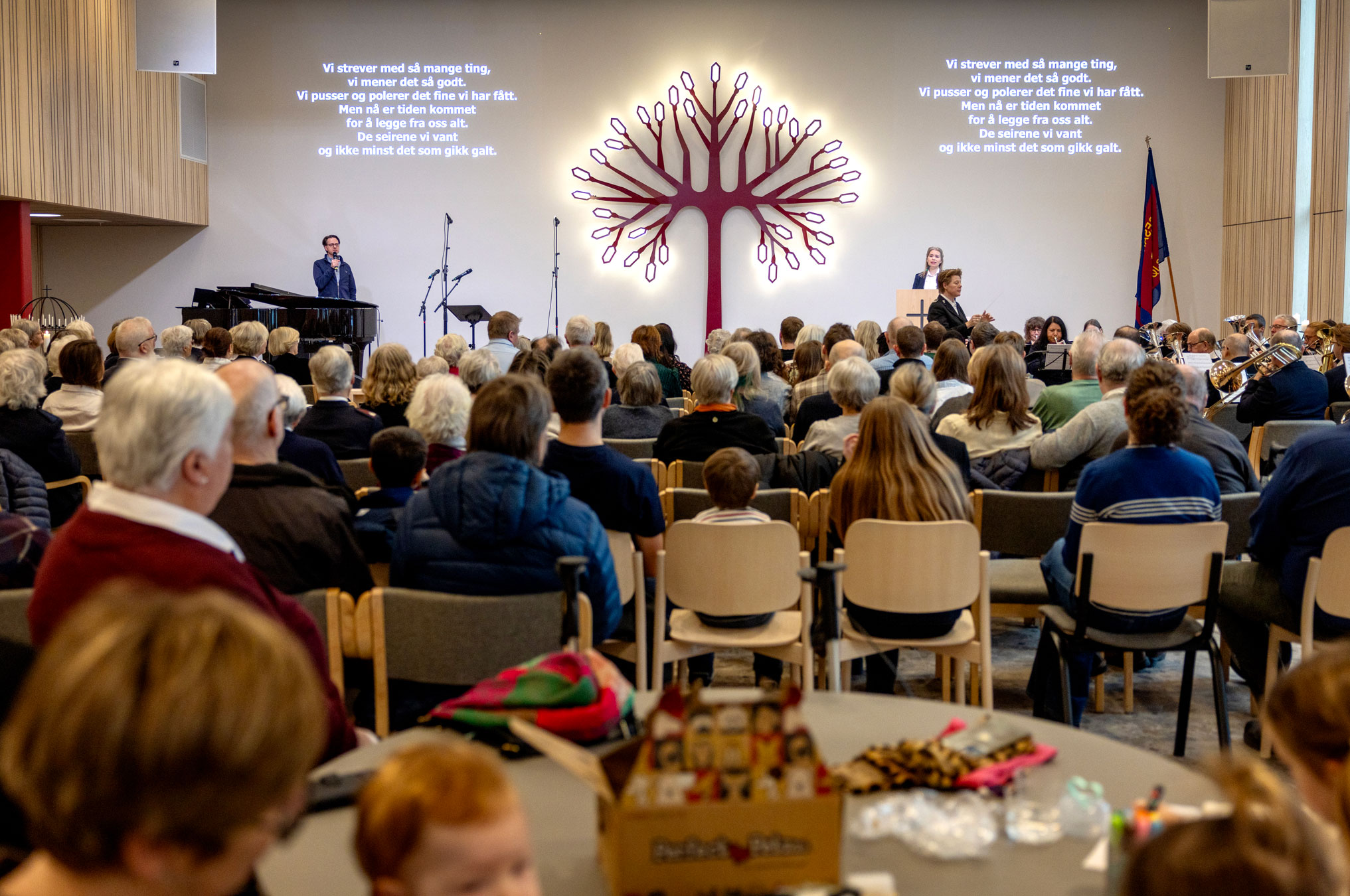 Foto: Fullsatt sal i et moderne kirkerom. I enden er det et kunstverk i form av et rødt metall-tre med led-lys i stedet for blader.