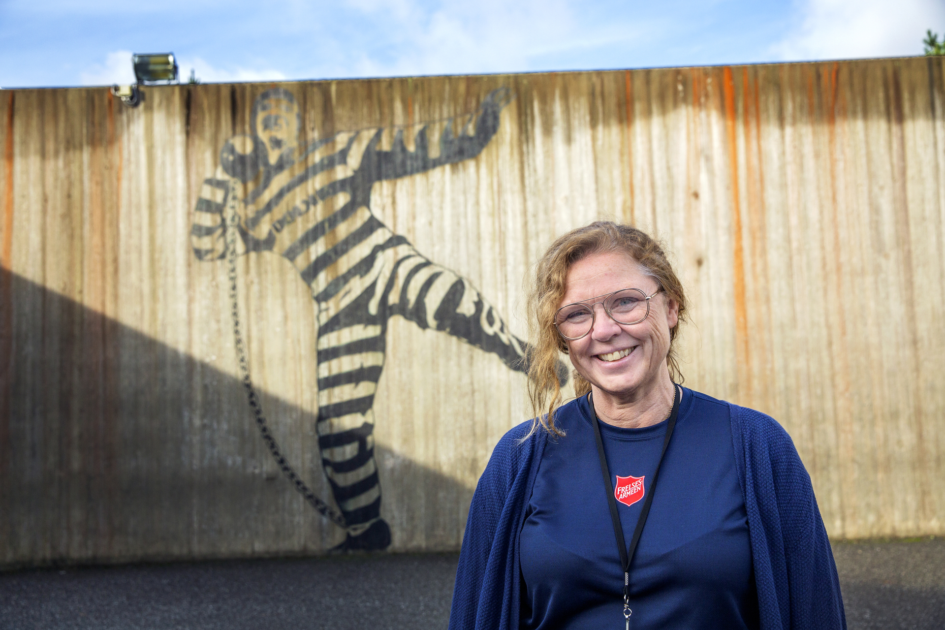 Foto: Dame i 50-årene med briller og blå klær med logo fra Frelsesarmeen som står foran en høy fengselsmur med grafitti av en mann i svart og hvitstripet fangedrakt. Han kaster en kule lenket til foten.