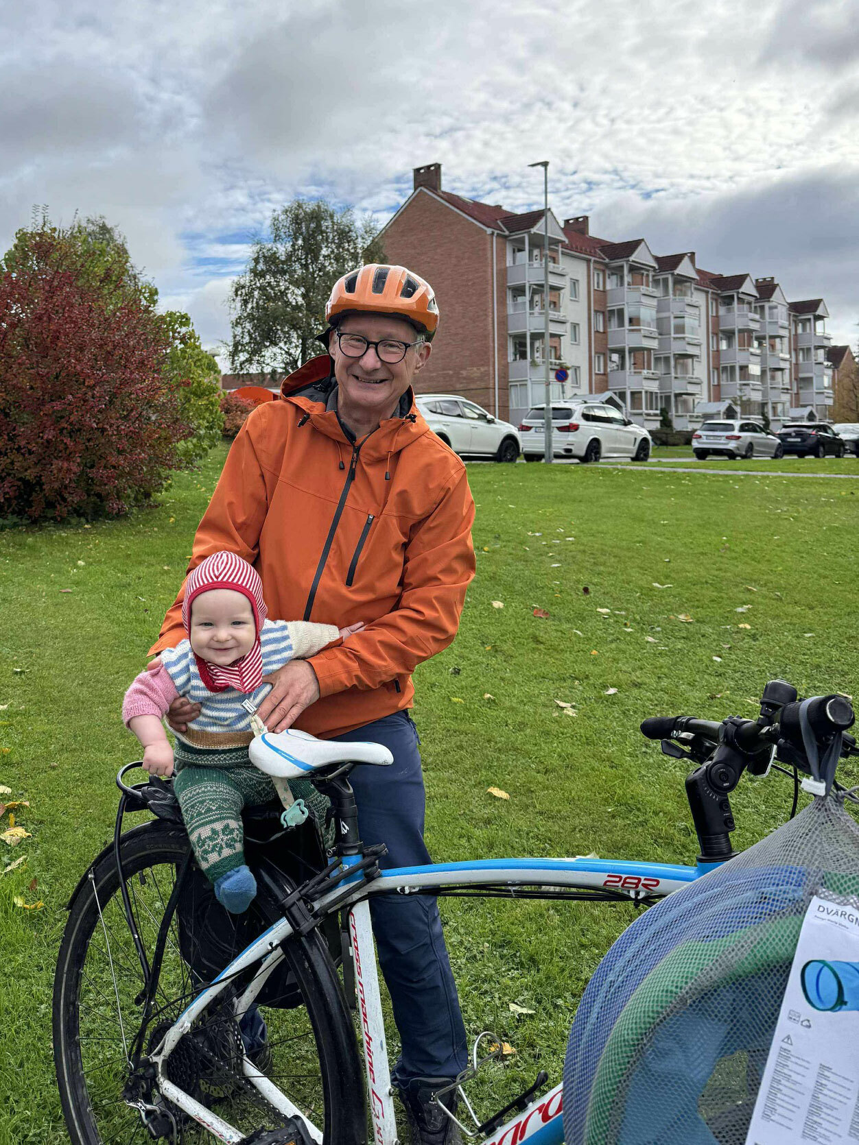 Foto: Mann i 70-årene med oransje hjelm og jakke holder en baby som liksom sitter bakpå sykkelsetet hans.