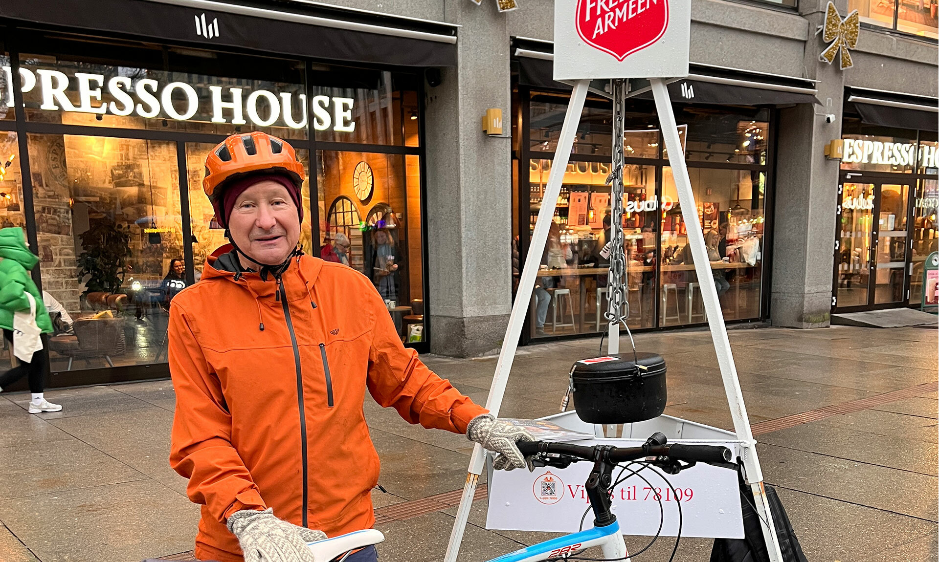 Foto: Eldre mann i oransje jakke og hjelp holder i en sykkel. Han står foran et stativ med Frelsesarmeens julegryte hengende ned i en kjetting. Han står foran det som er et kjøpesenter med kafé på første plan.