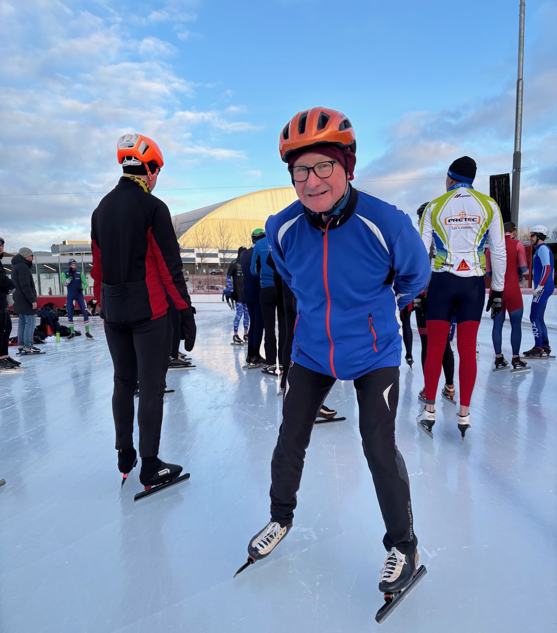 Foto: Mann i 70-årene med oransje hjelp står med skøyter på isen. Rundt er det flere folk.