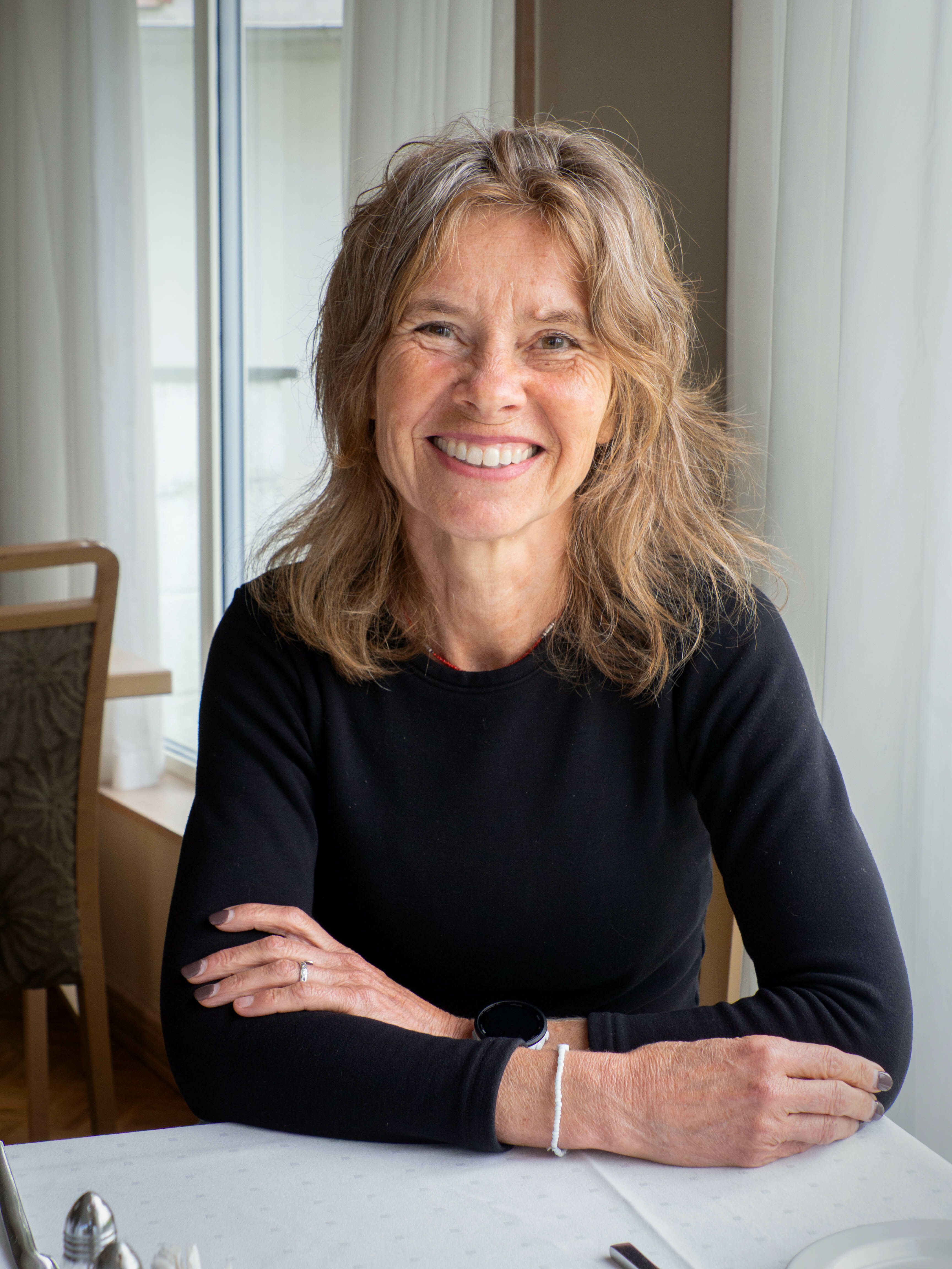 Foto: En kvinne med lyst, langt, litt bølgete hår sitter ved et bord og smiler mot kamera. Hun har på seg svart genser.