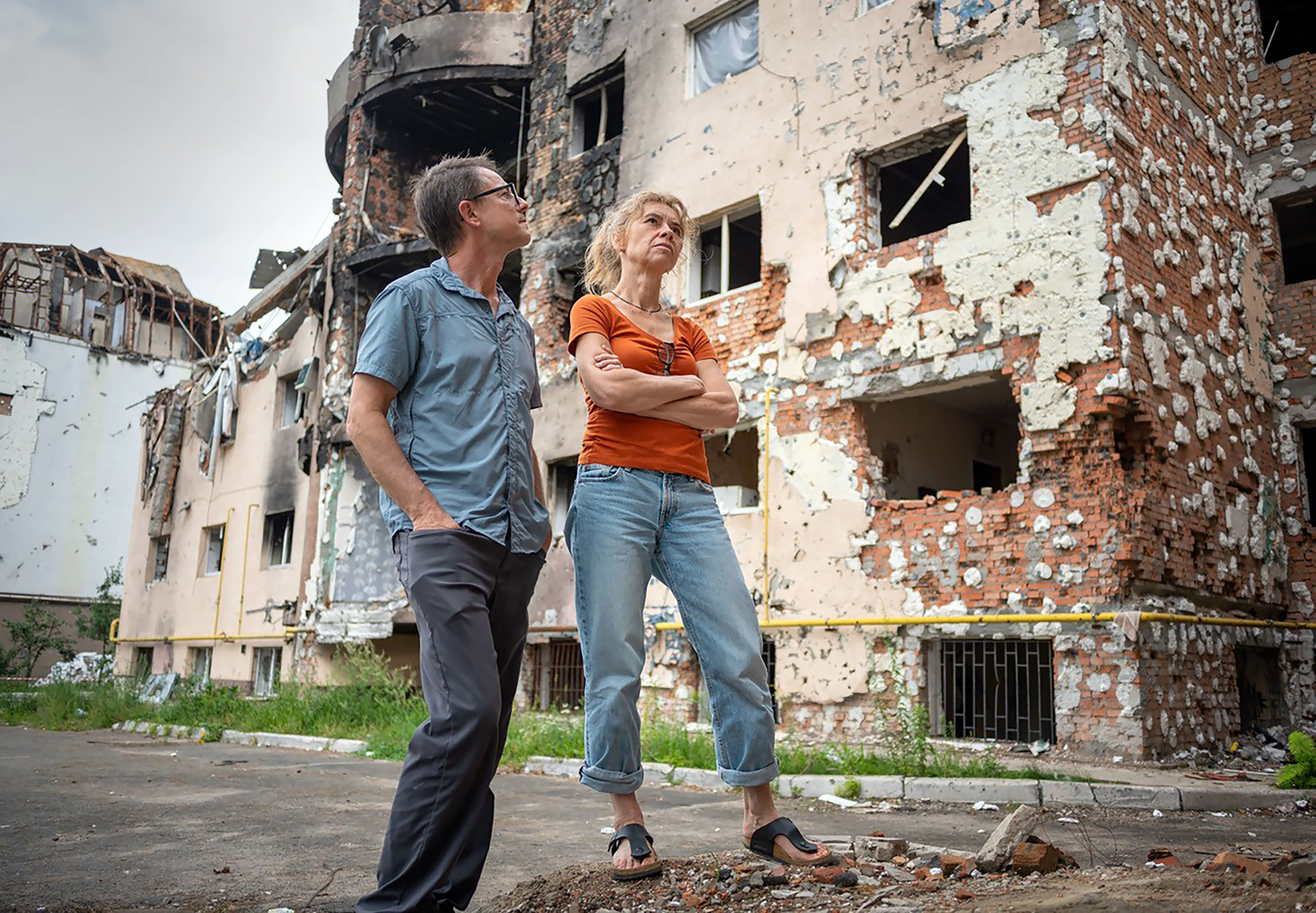 Foto. En kvinne og en mann står i en gate foran ødelagte murbygg. Bildet er tatte litt nedenfra, mannen står med tennene i lomma og ser opp mot bygget, kvinnen har armene i kors og ser oppover med et alvorlig uttrykk.