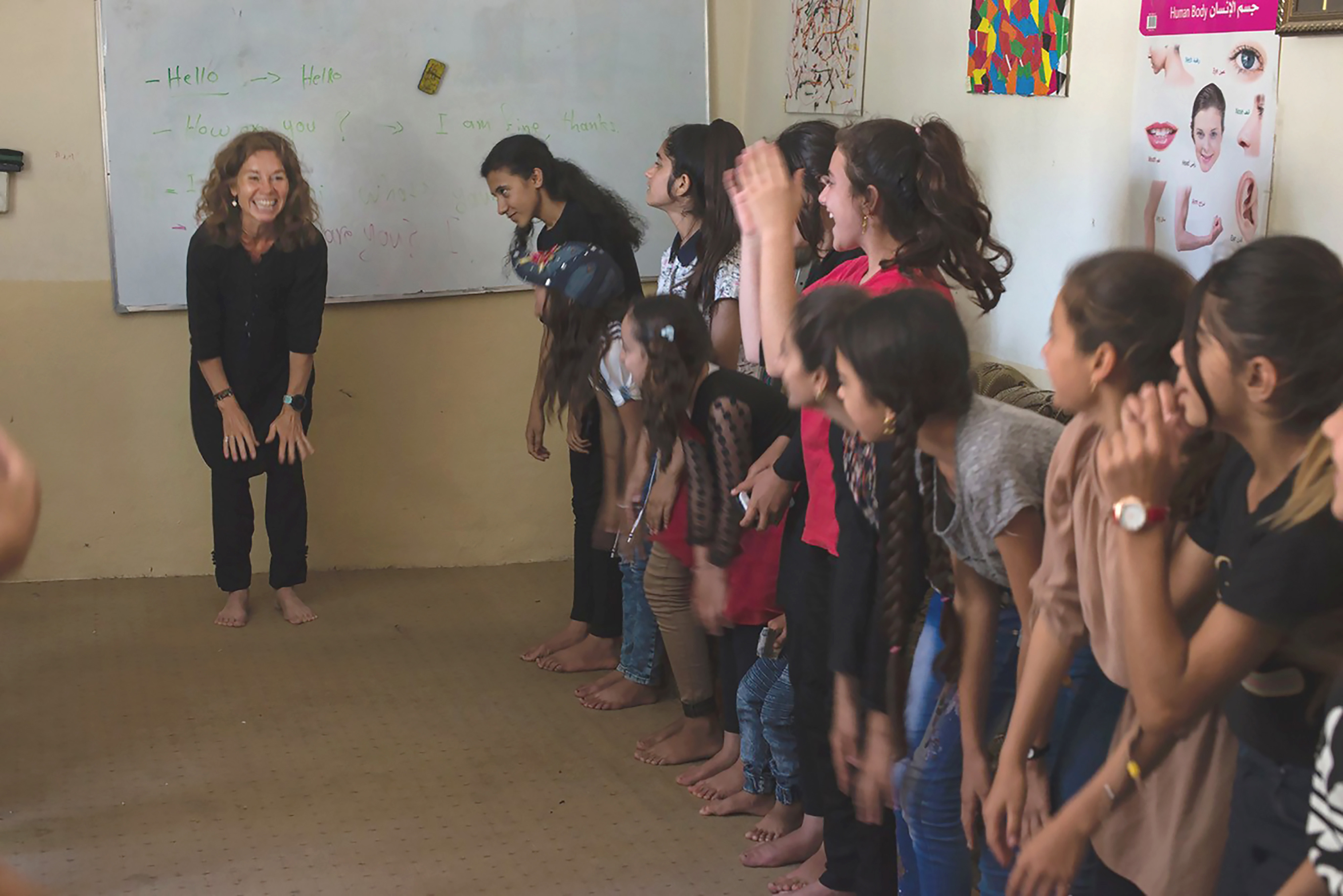 Foto: Enk kvinne i svarte klær står og smiler og holder seg på knærne. Langs veggen til høyre for henne står en rekke barn og ser på henne og smiler.
